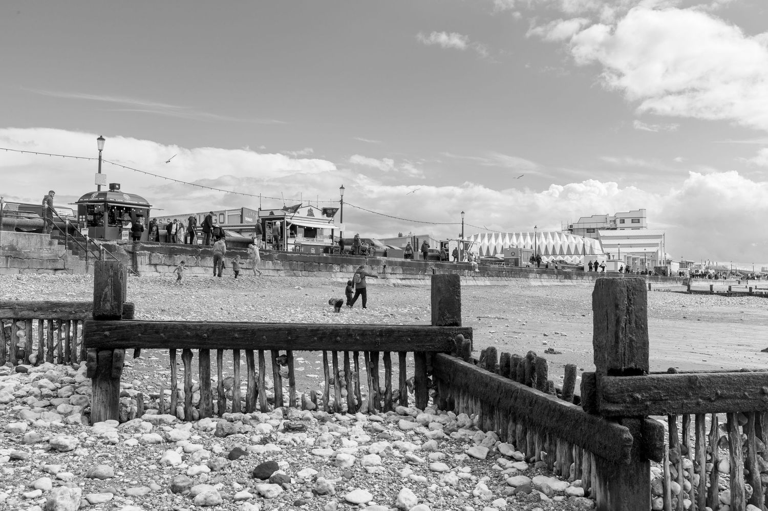 Panoramic black and white view of a wooden pier with posts stretching into calm water under cloudy skies.