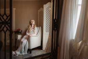 A person in a white dress stands in a bright room with lattice panels and sheer curtains.