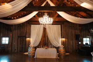 Elegant barn wedding venue interior with white fabric draping, crystal chandeliers, and rustic wooden beams.
