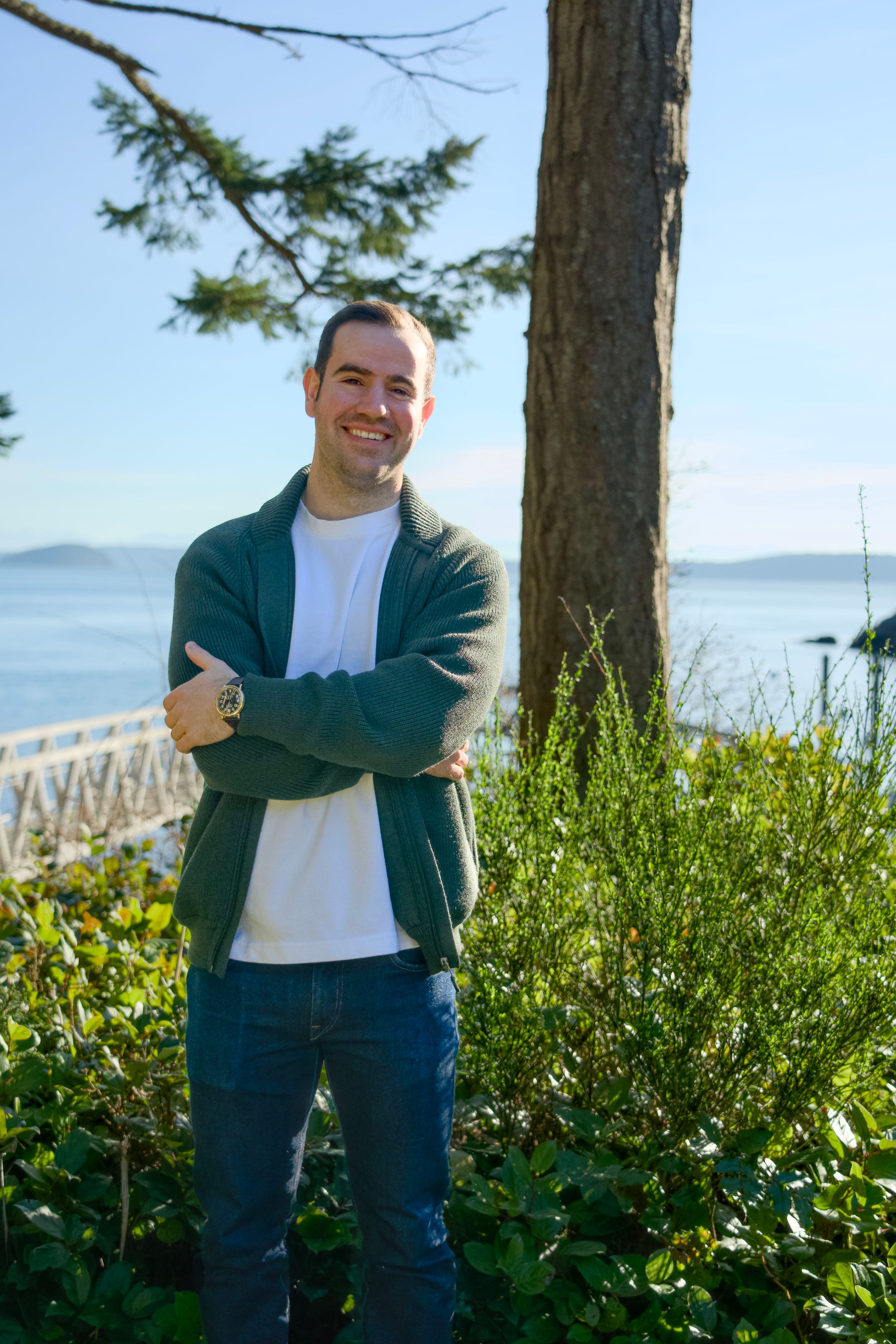 Seattle photographer, Mathew Grosky, posing with arms crossed and smiling in front of a scenic coastal backdrop.