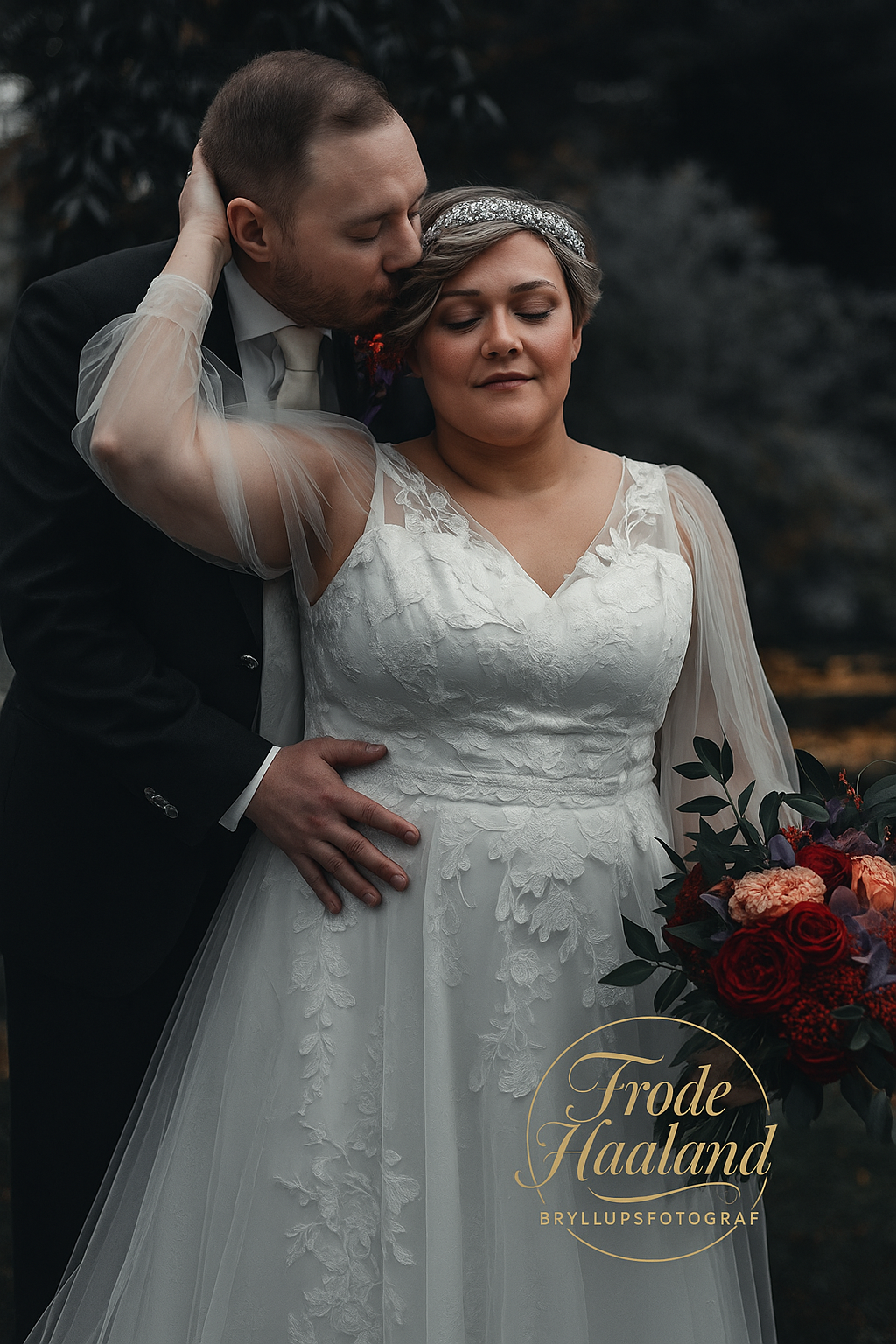 Groom tenderly kissing bride on cheek as she holds a red bouquet in a lace wedding gown.