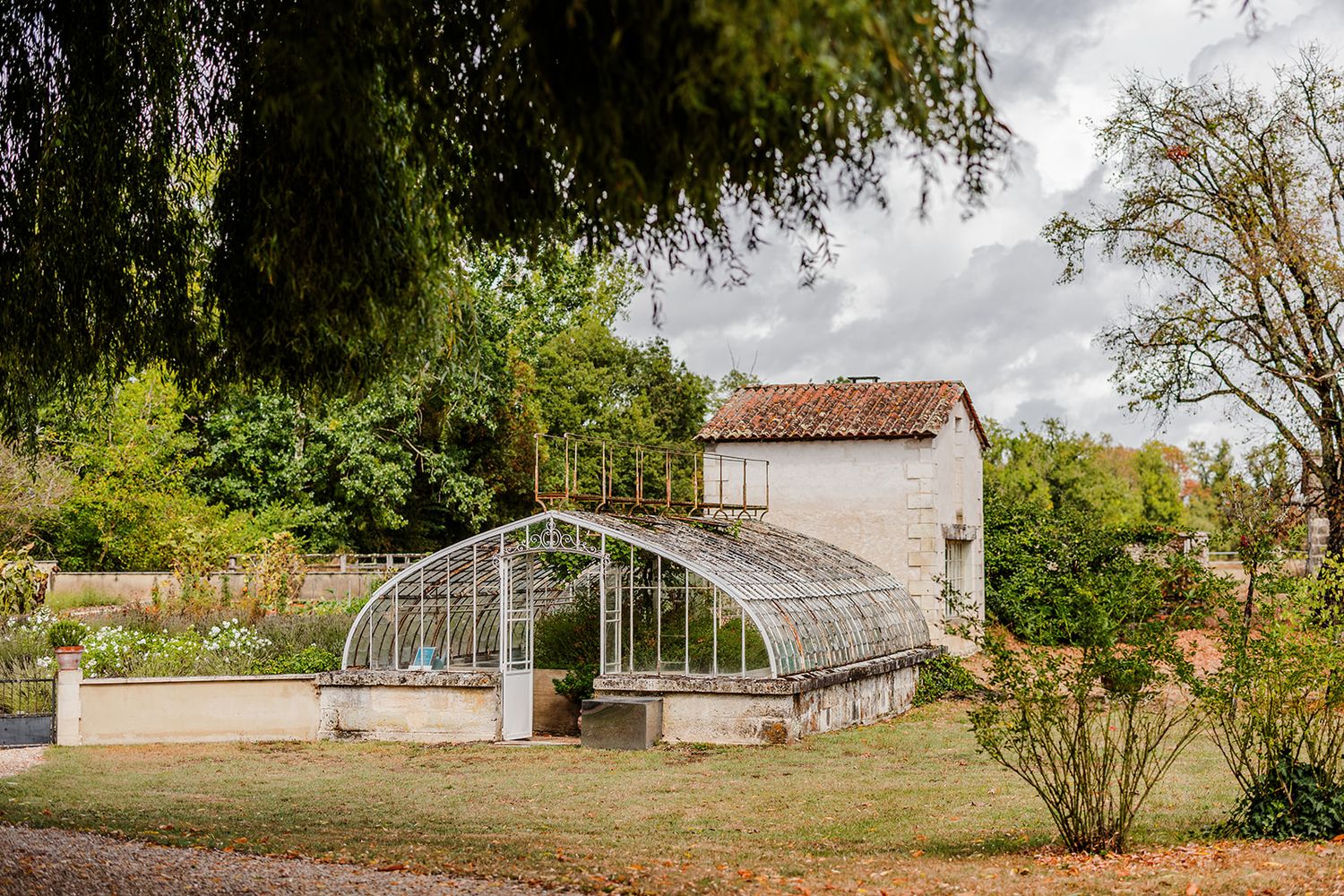 A series of old greenhouses and farm buildings stand in a row along a rural property under cloudy skies.