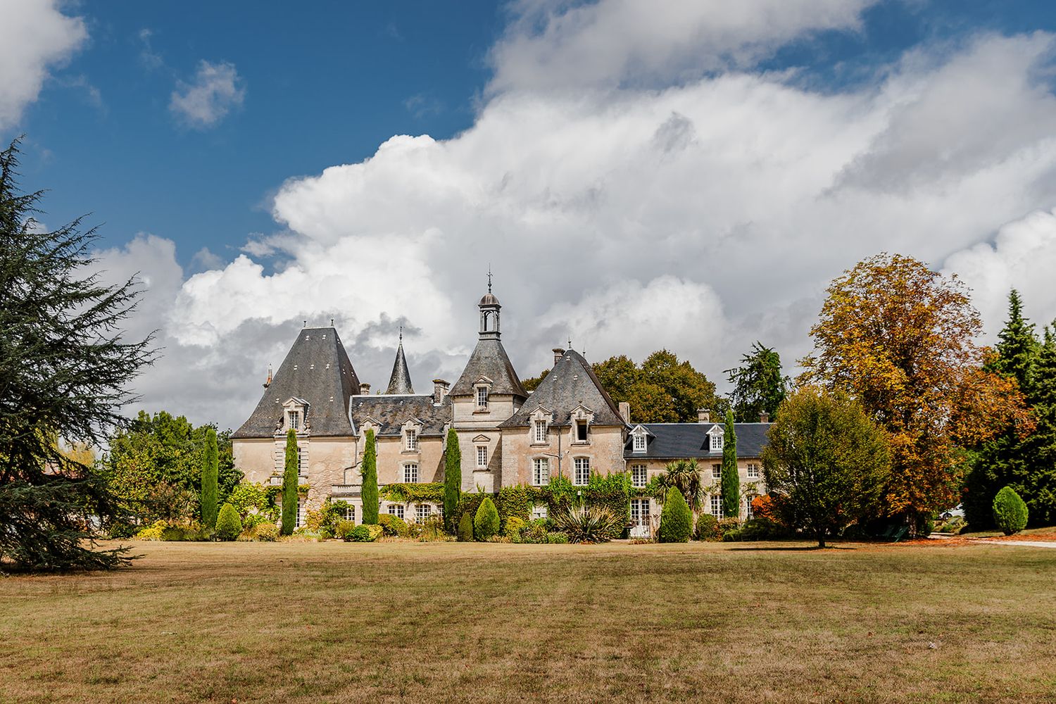 A panoramic view of a historic stone manor house with multiple buildings set against a blue sky with white clouds.