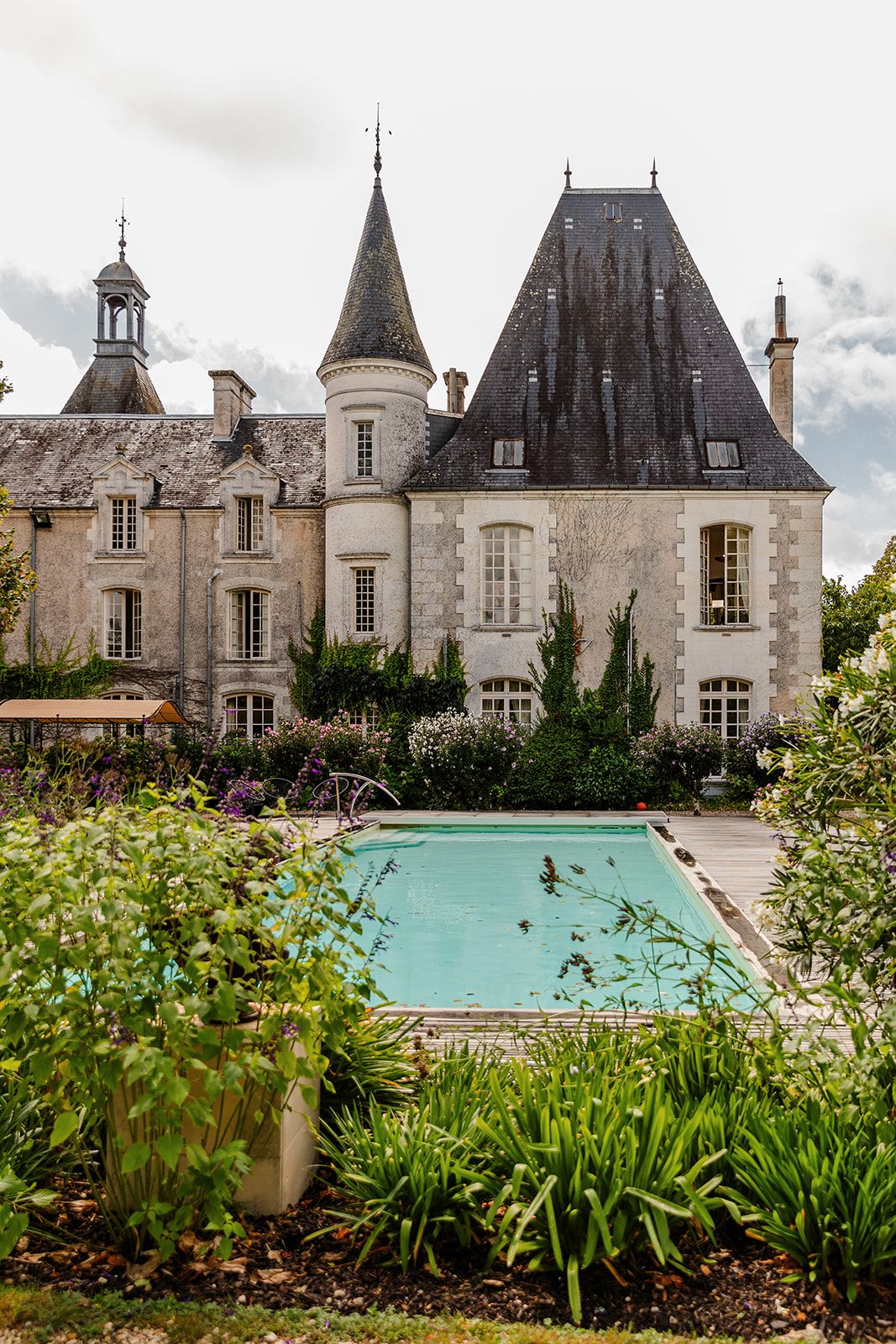 A panoramic view of a French chateau with a turquoise swimming pool surrounded by manicured gardens and landscaping.