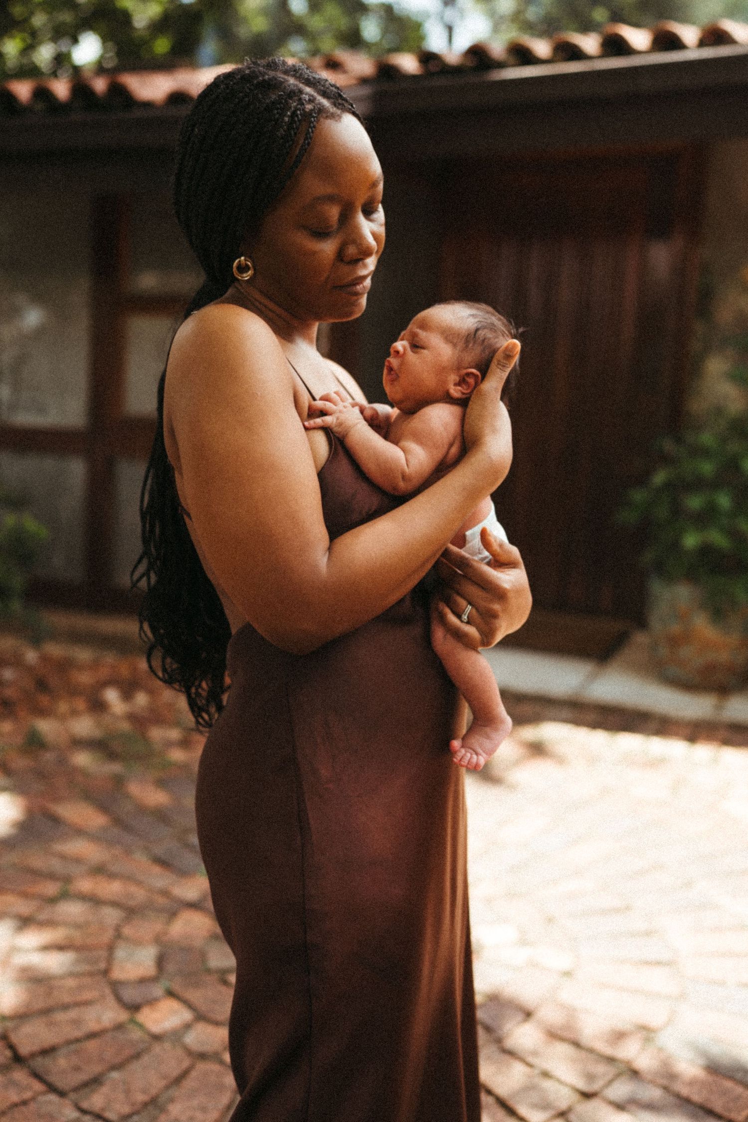 Mother in brown dress holding newborn baby in outdoor sunlit setting.