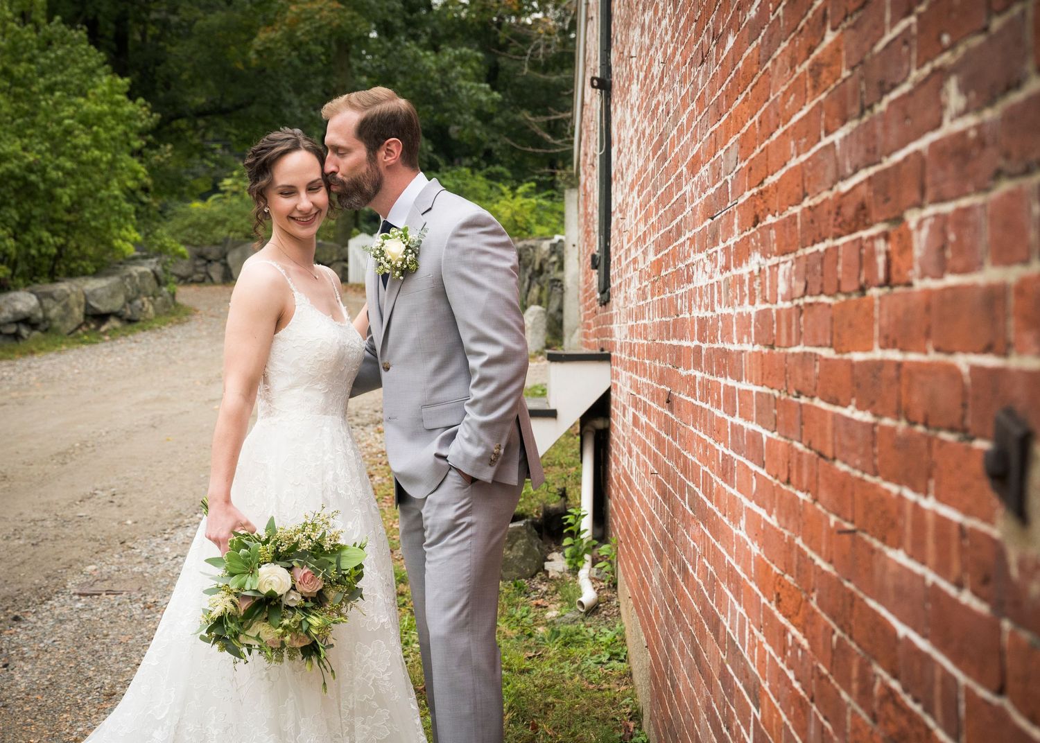 A couple in wedding attire shares a romantic moment against a rustic brick wall backdrop.