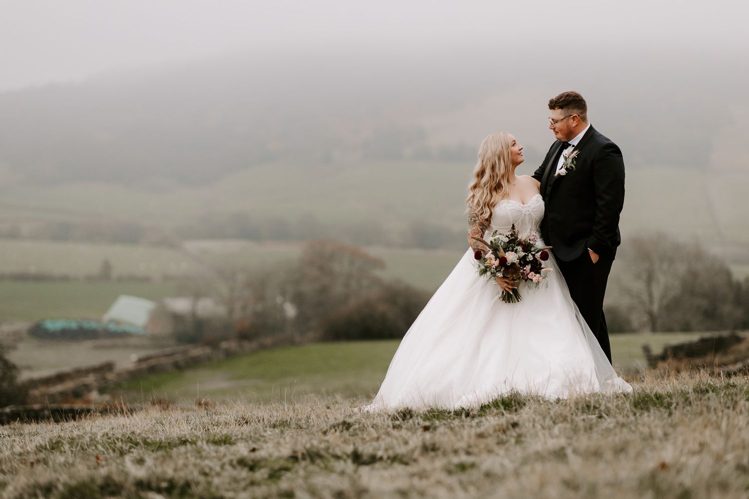 A couple in wedding attire walks through misty hills and fields during an overcast day in the countryside.