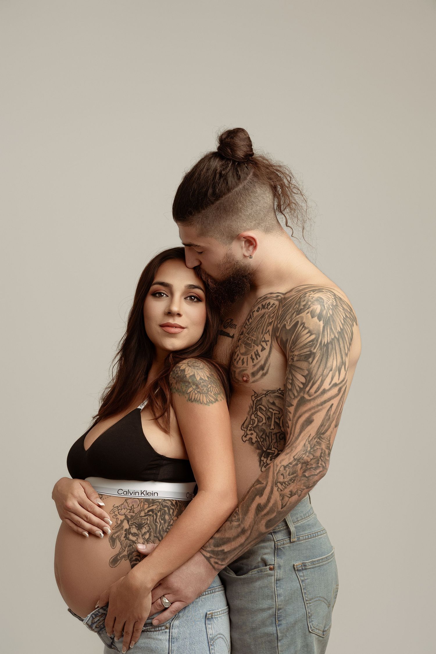 Intimate maternity photo showing couple embracing against neutral background, with visible tattoos and casual attire. Paloma Schell