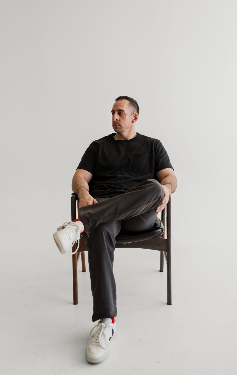 Person in black t-shirt and gray pants sitting casually on wooden chair against white background.