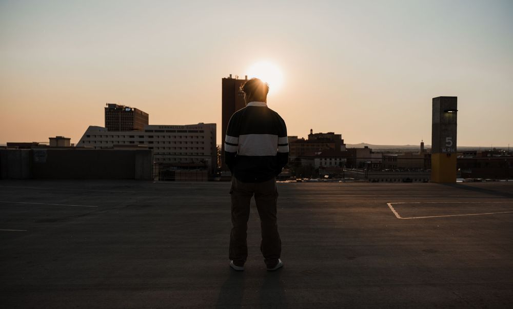 A series of silhouetted figures walking against a sunset sky in an urban rooftop setting.