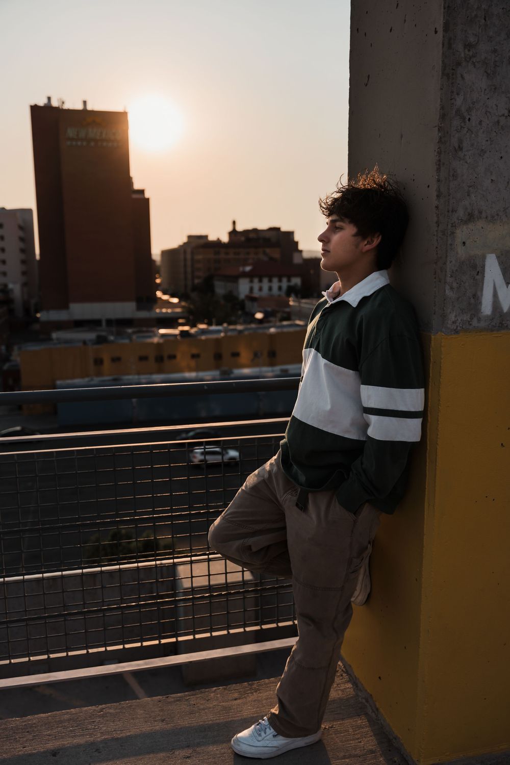 Person in casual wear sits on a rooftop ledge at sunset with city buildings in the background.