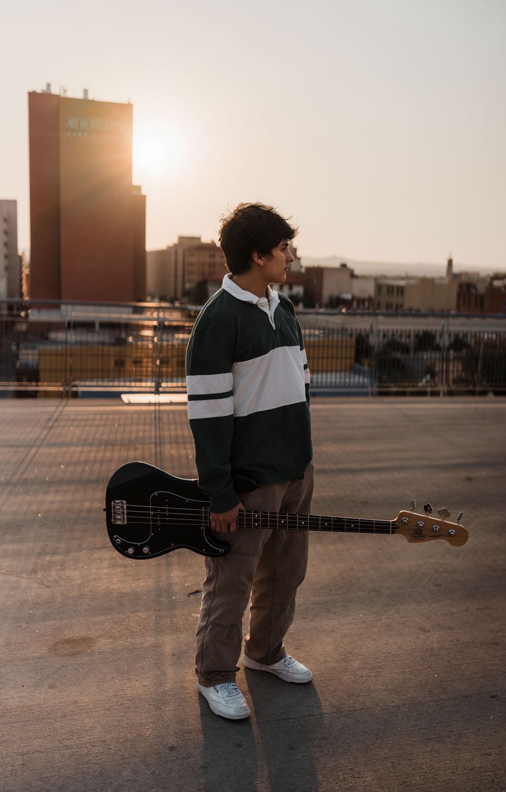 Musician stands on urban rooftop at sunset holding bass guitar in silhouette against golden sky.