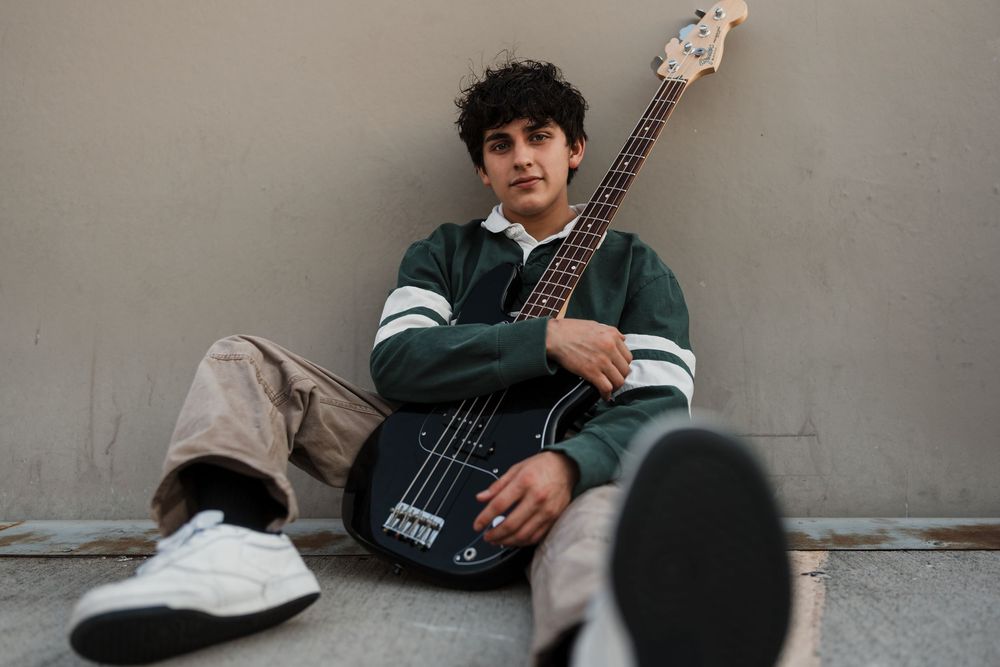 A person wearing a green jacket plays an electric bass guitar while sitting casually against a gray wall.