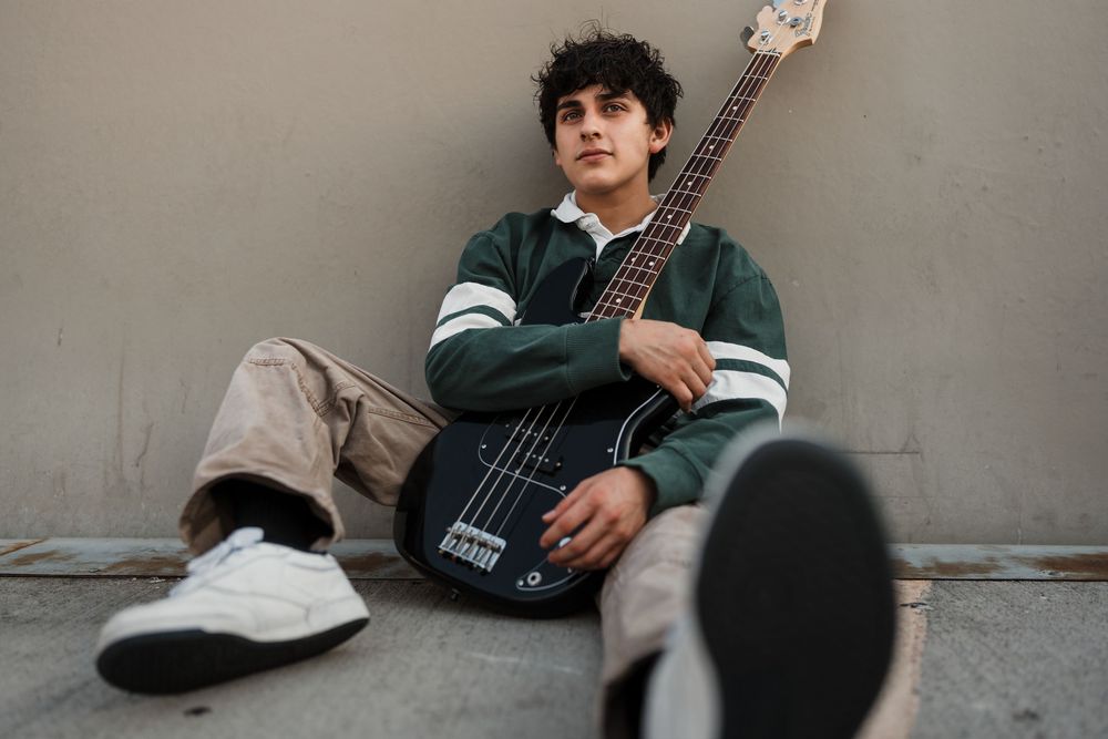 A person in casual attire sits on the ground against a wall while holding a bass guitar.