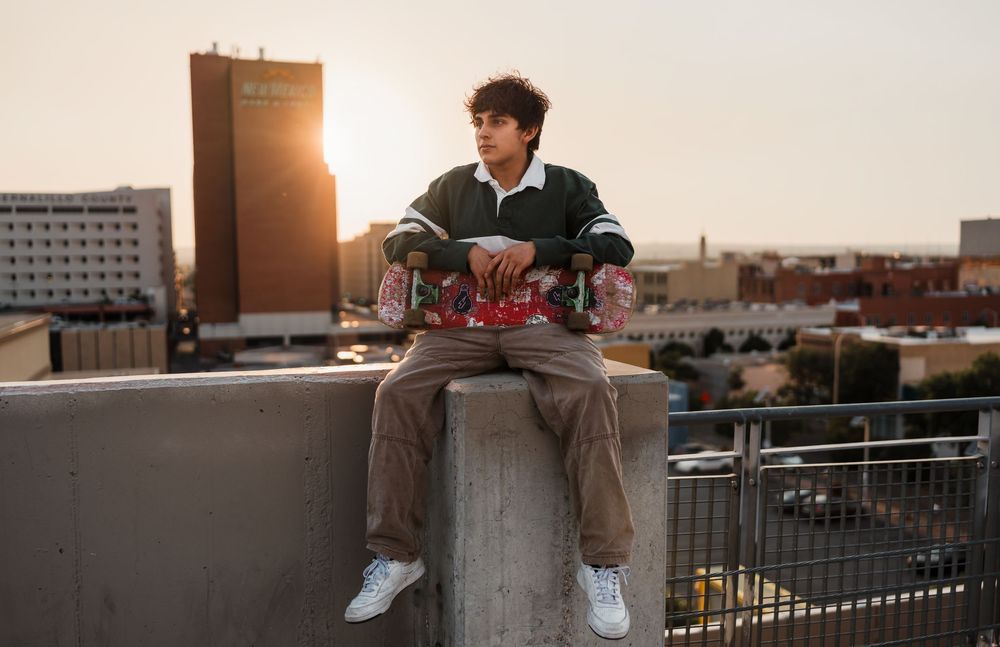 Person sitting on rooftop edge with skateboard at sunset against urban cityscape backdrop.