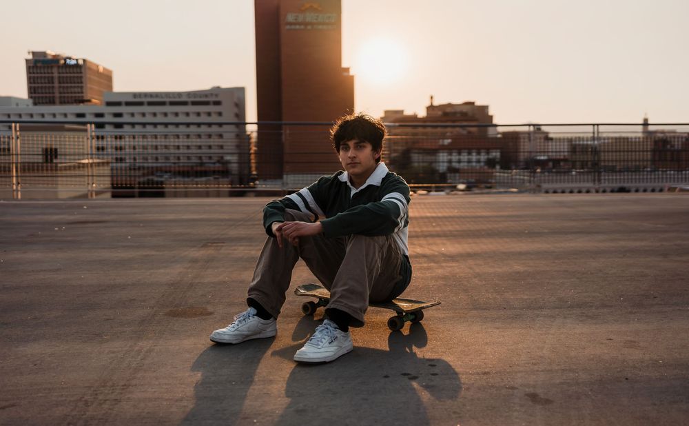 Person in dark clothing sits on an urban rooftop at sunset with cityscape views in the background.