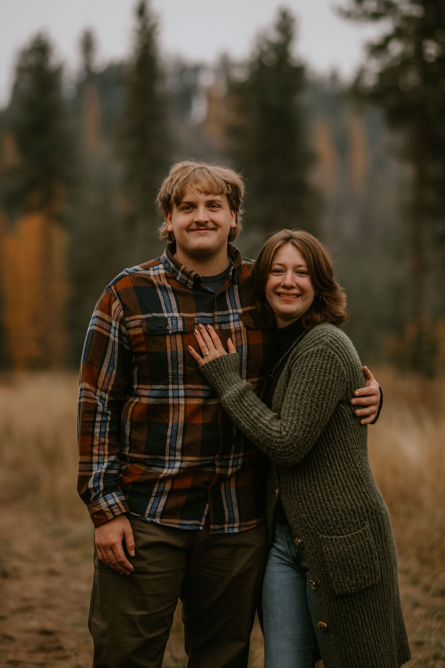 A couple in autumn attire laughs and embraces while standing in a field with evergreen trees in the background.