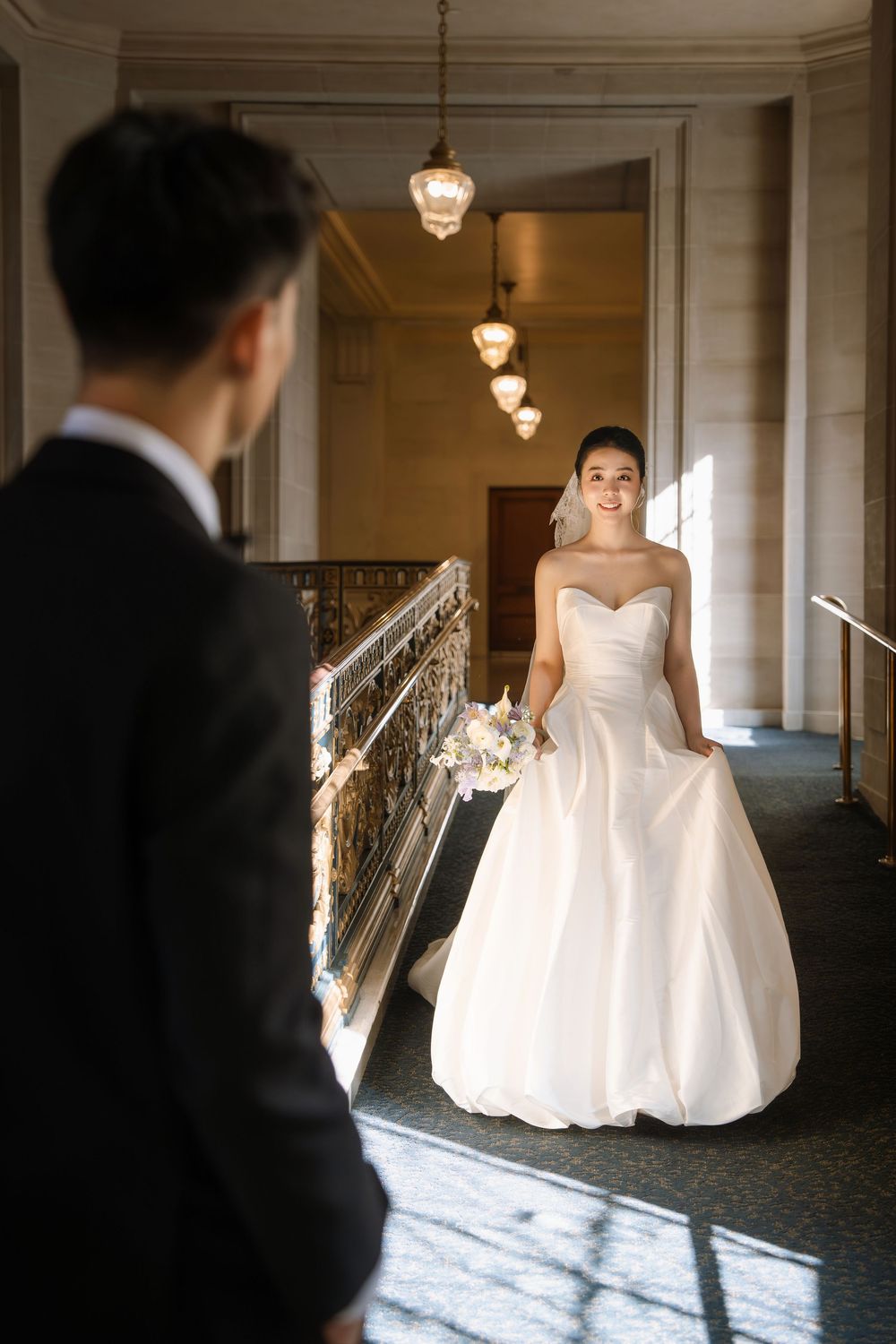 Wedding couple in elegant attire photographed in a grand hallway with pendant lighting and dramatic shadows.