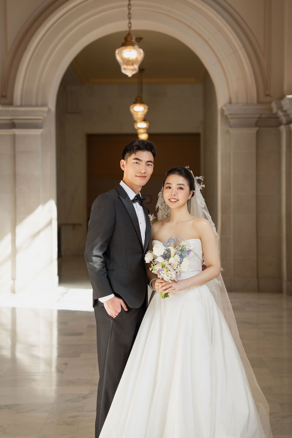 A couple in formal wedding attire poses in an elegant hallway with arched columns and pendant lighting.