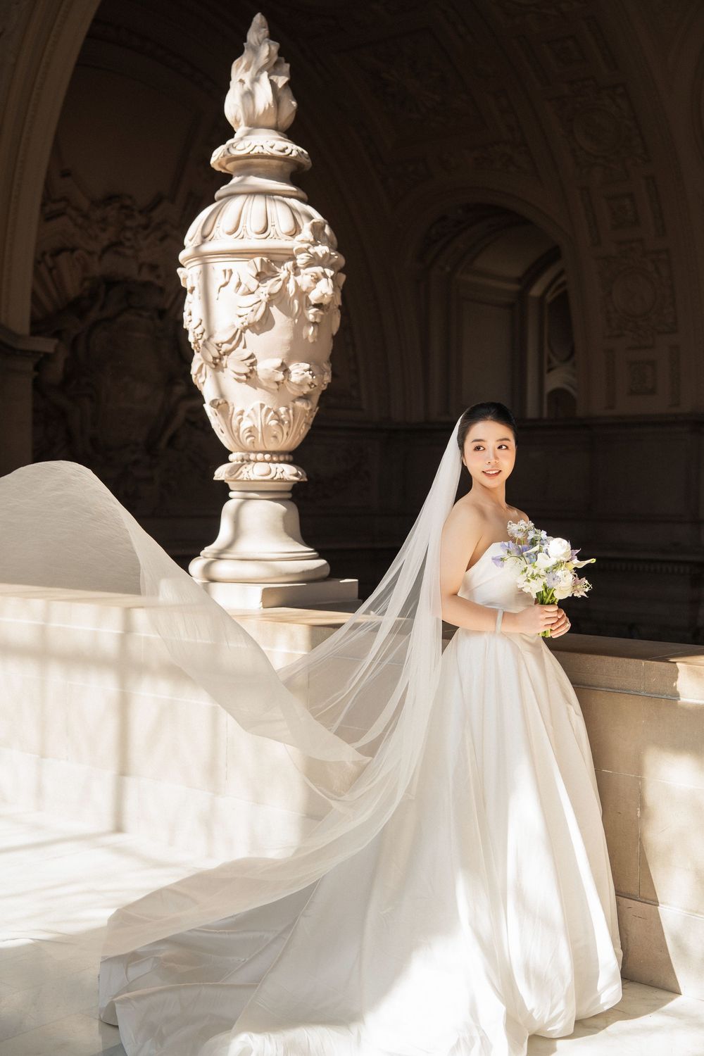 Elegant bride in flowing white gown and veil poses gracefully against ornate architectural columns in natural light.