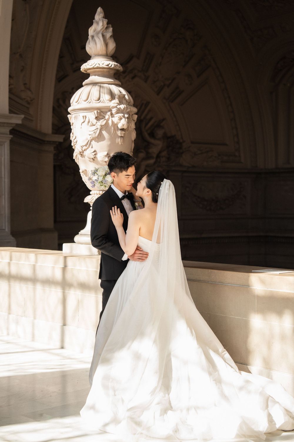 A bride in a flowing white veil poses in an elegant architectural setting with ornate stone columns and dramatic lighting.