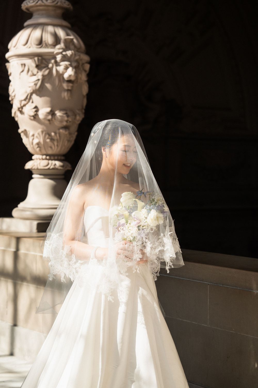 A bride in a flowing white wedding dress and veil stands elegantly against a stone column backdrop.