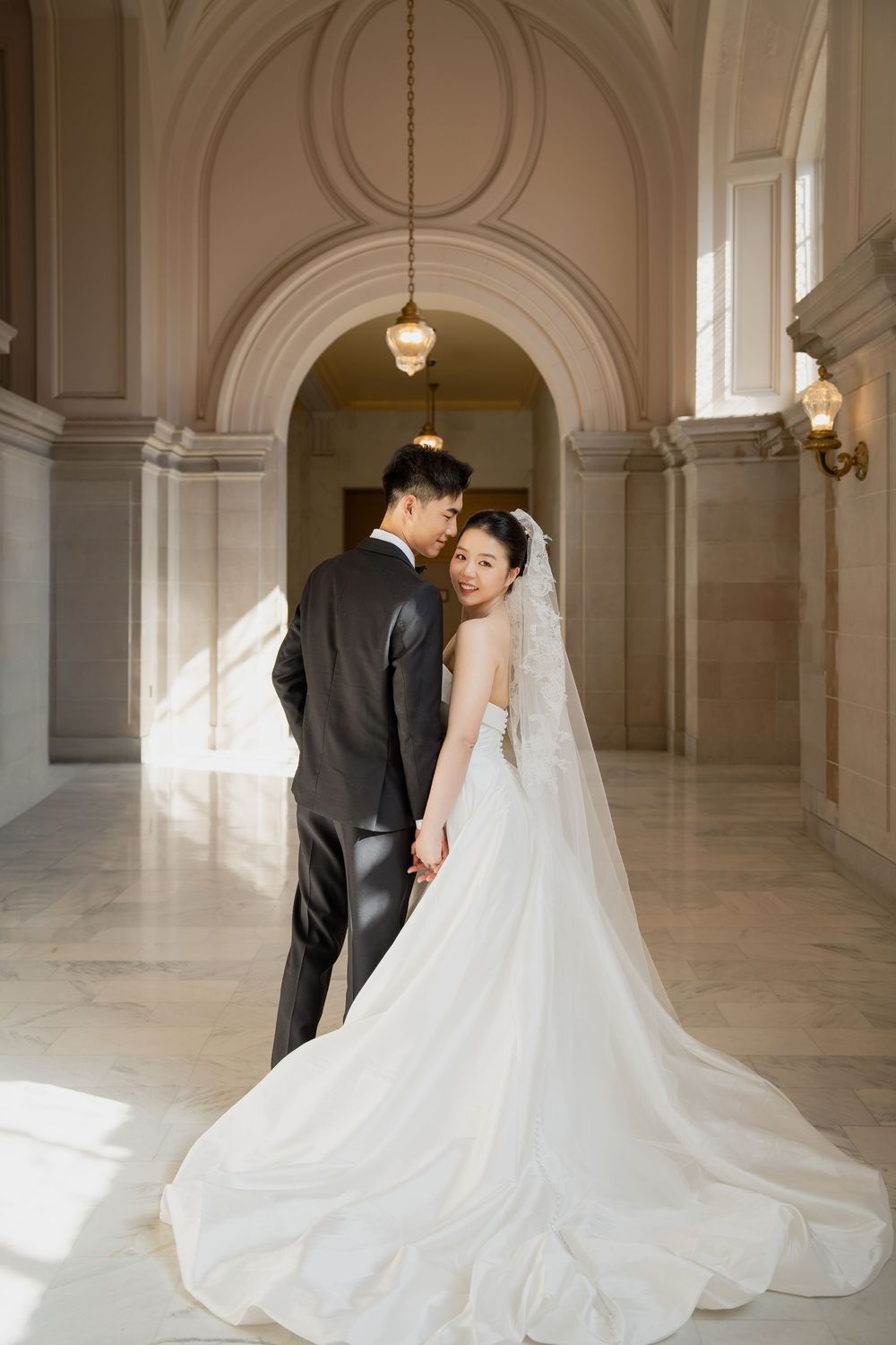 An elegant wedding couple embraces in a grand marble hallway with arched ceilings and classical architecture.