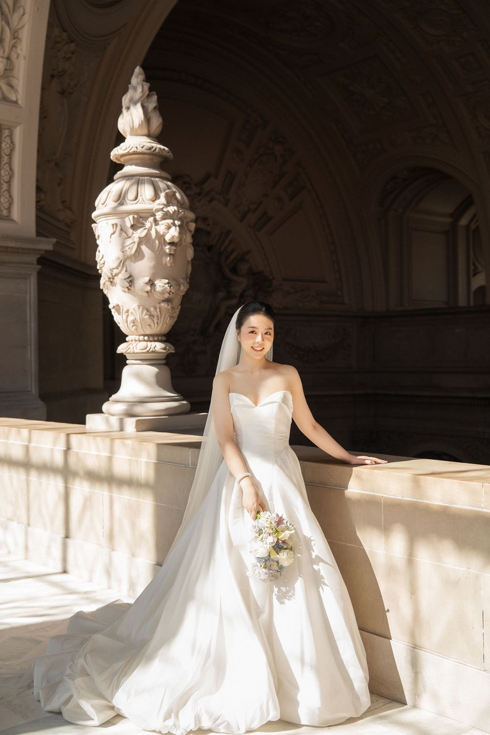 A bride in a white wedding gown sits gracefully in an elegant architectural setting with ornate columns and natural light.