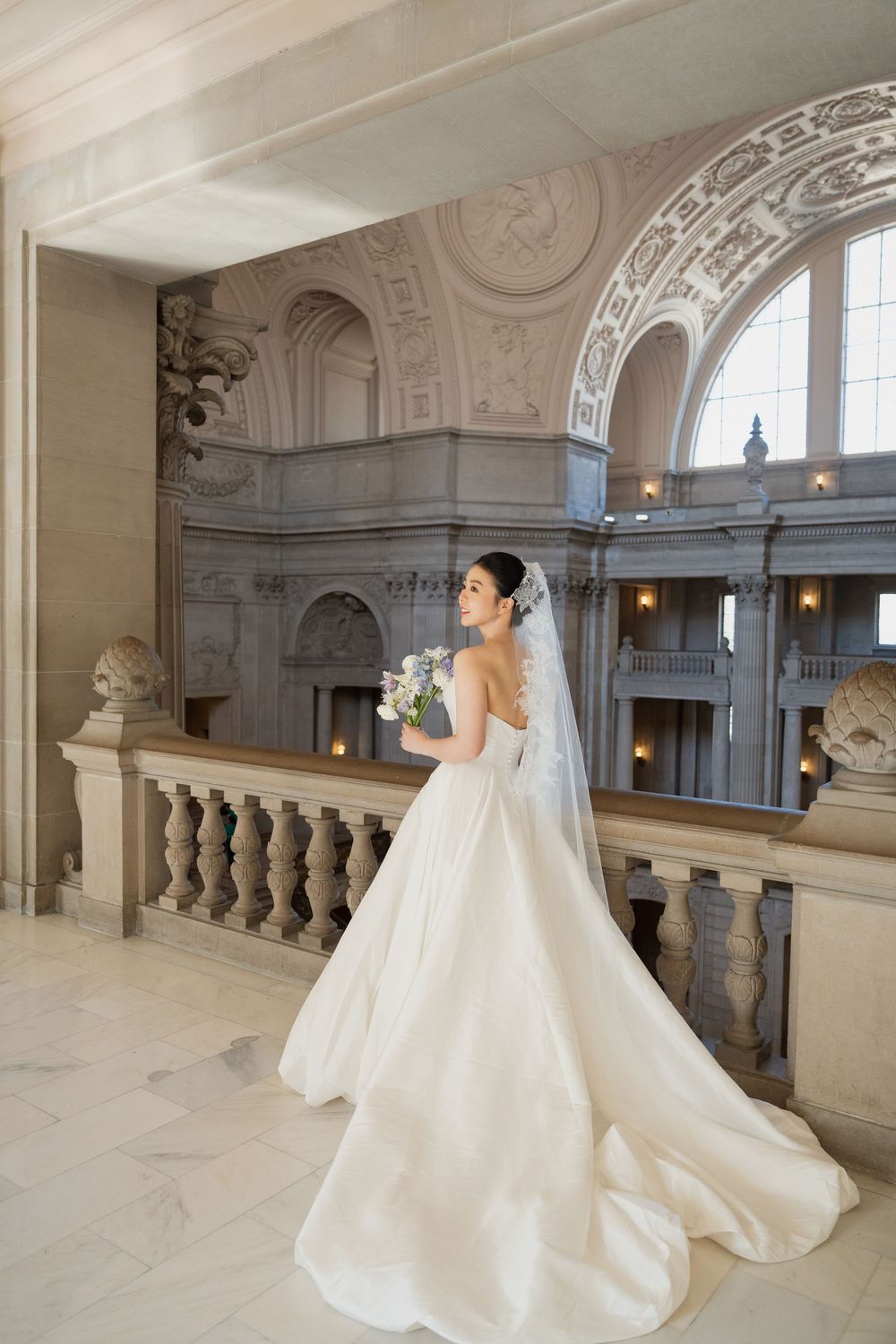 Elegant bride in flowing white gown poses in ornate marble hallway with classical architectural details and natural lighting.