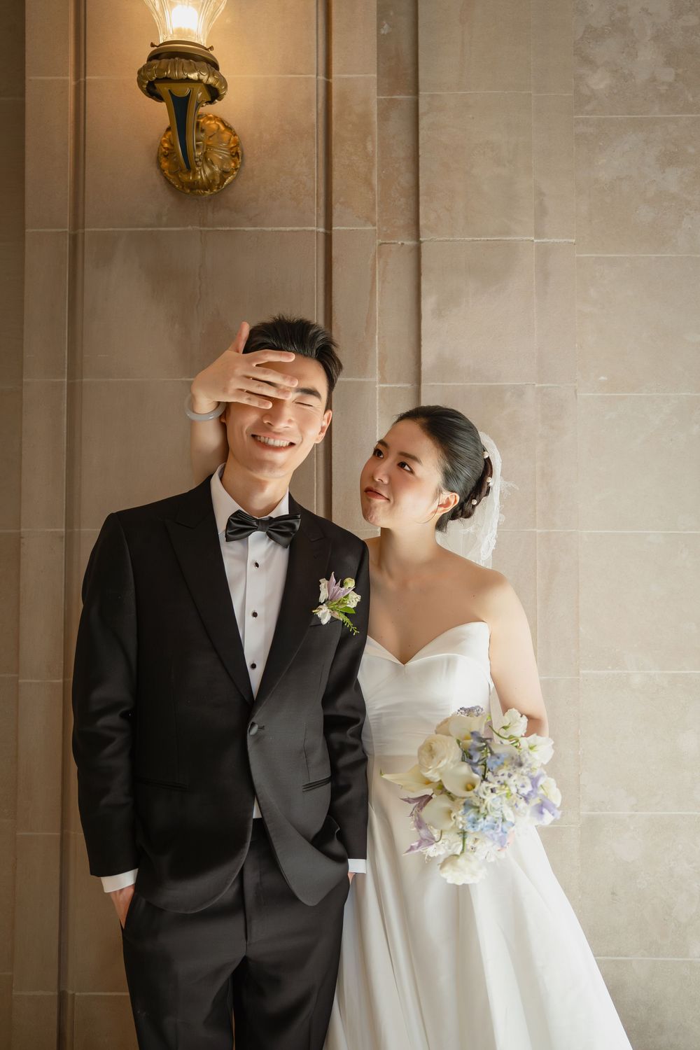 A joyful couple shares playful moments in formal wedding attire against a neutral stone wall with sconces.