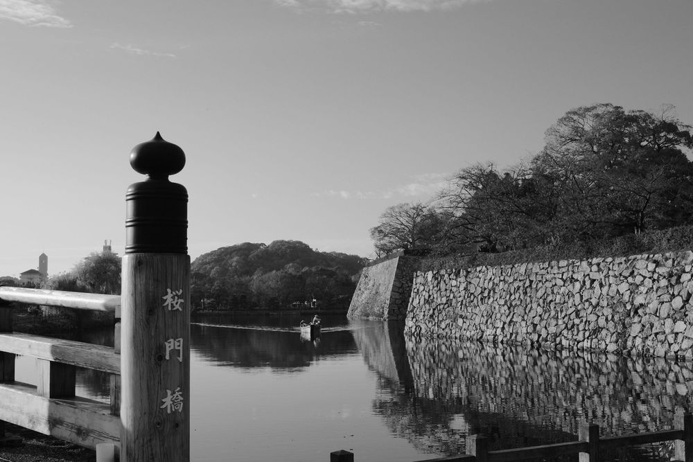 Black and white panoramic view of a stone wall stretching along a hillside with trees and buildings in the background.
