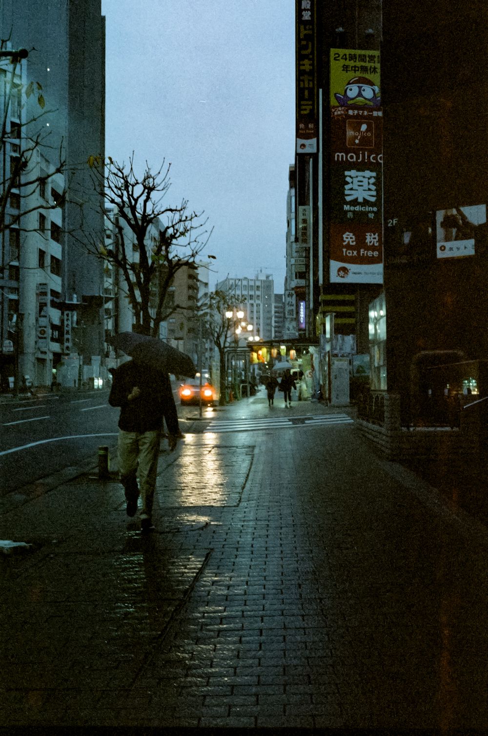 Rainy evening city street with silhouetted figures walking under umbrellas against glowing urban lights.