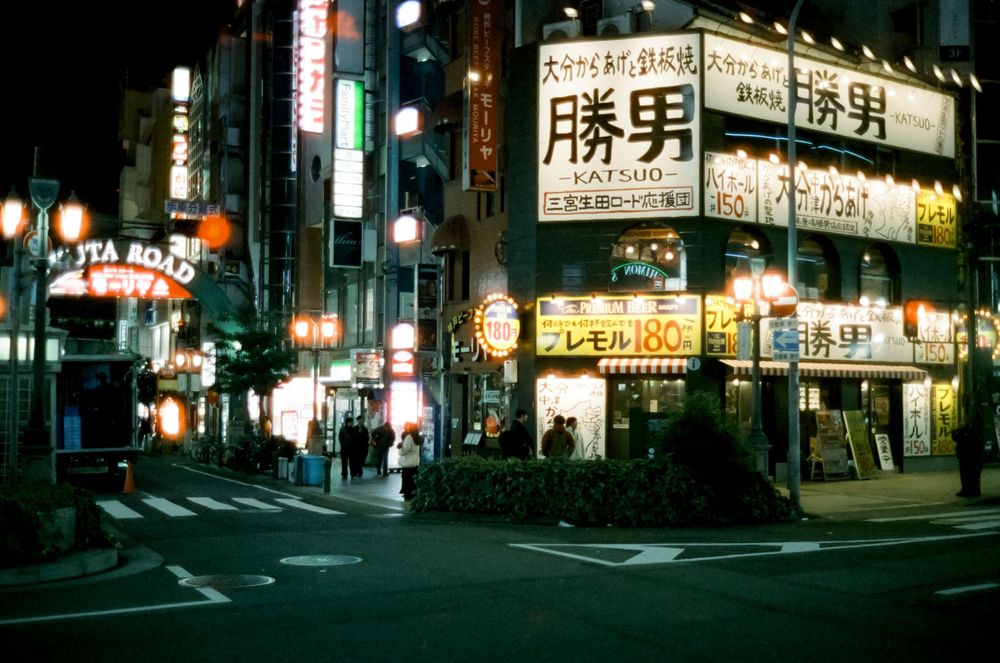 Illuminated Japanese street signs and storefronts glow against the dark night sky in an urban neighborhood.