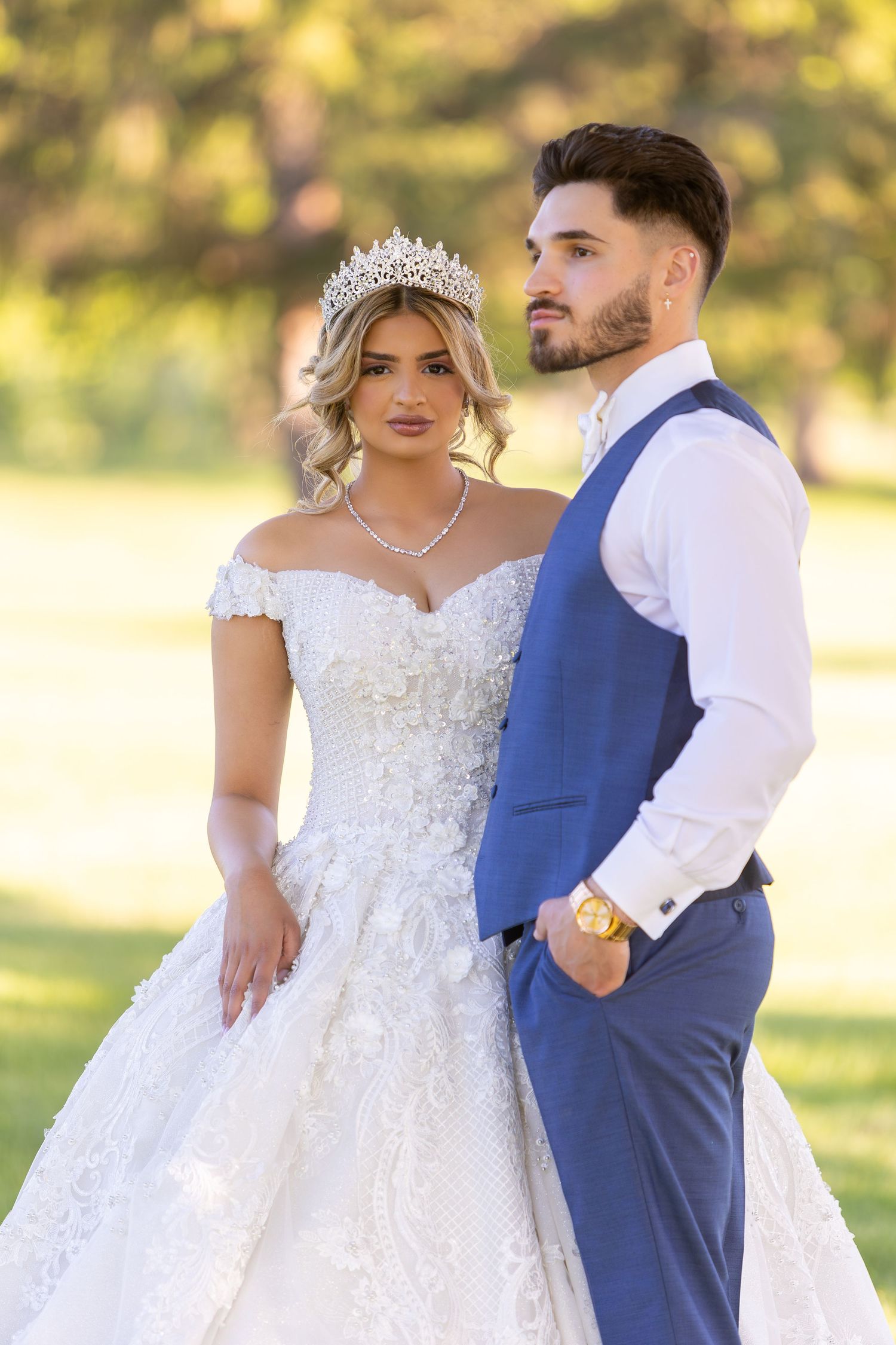 A bride in a lace off-shoulder gown and crystal crown poses with groom in navy vest outdoors during golden hour.