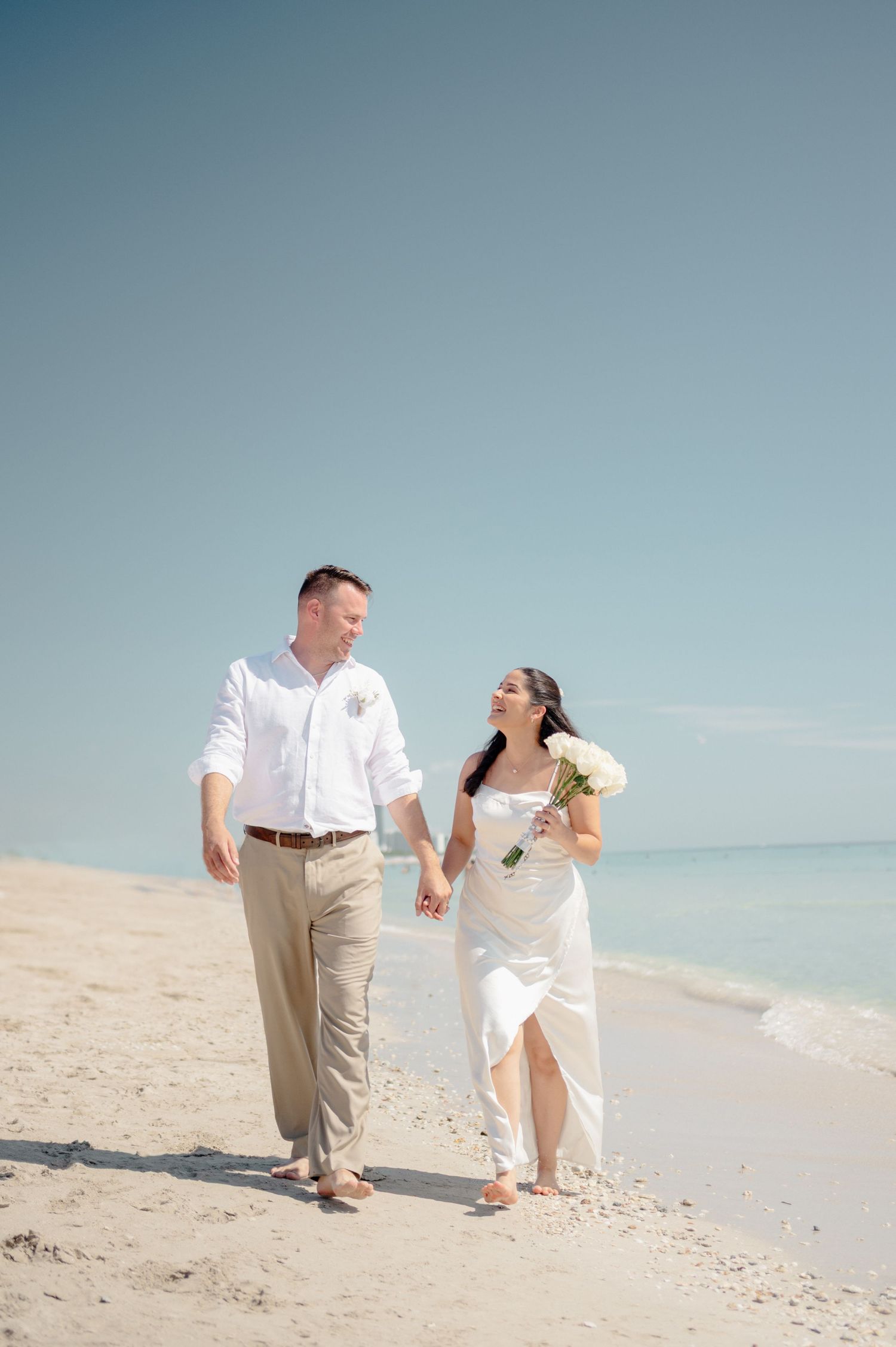 A couple strolls hand in hand along a pristine white sand beach with turquoise waters and clear blue skies.