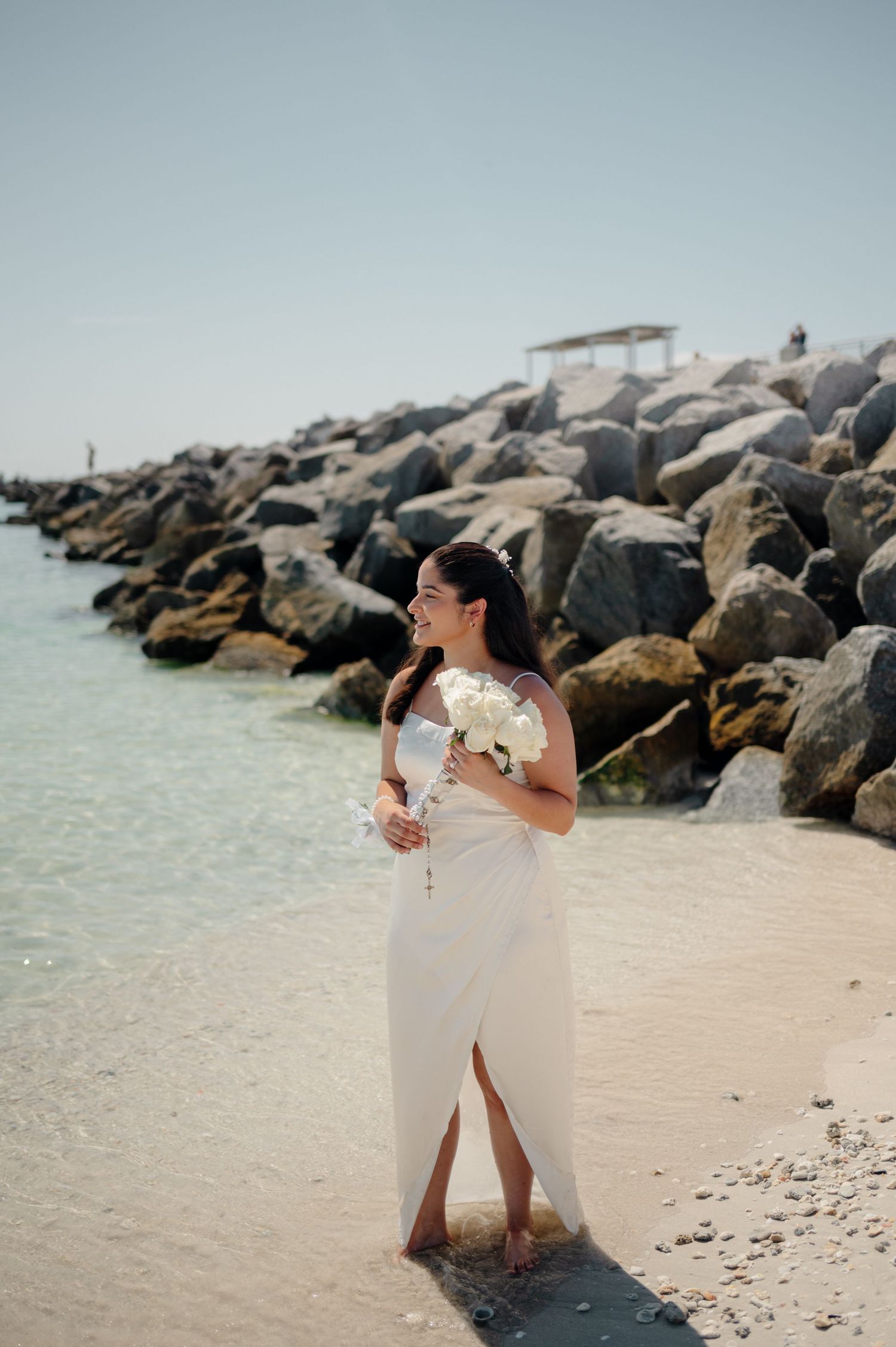 Two people in white dresses share a joyful moment on a beach with a rocky jetty in the background.