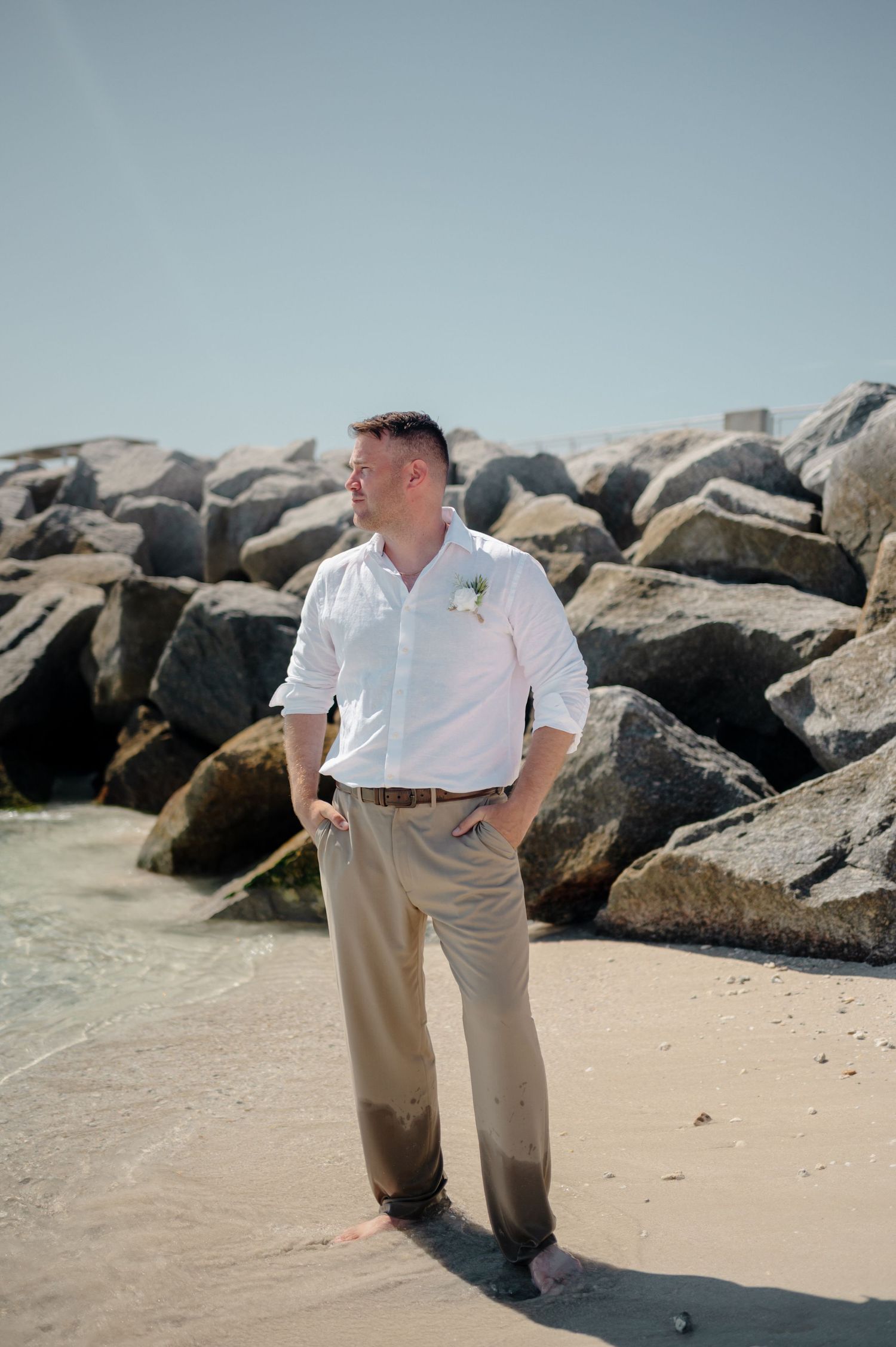A person in white shirt and khaki pants stands on a sandy beach near large rocky breakwater boulders.