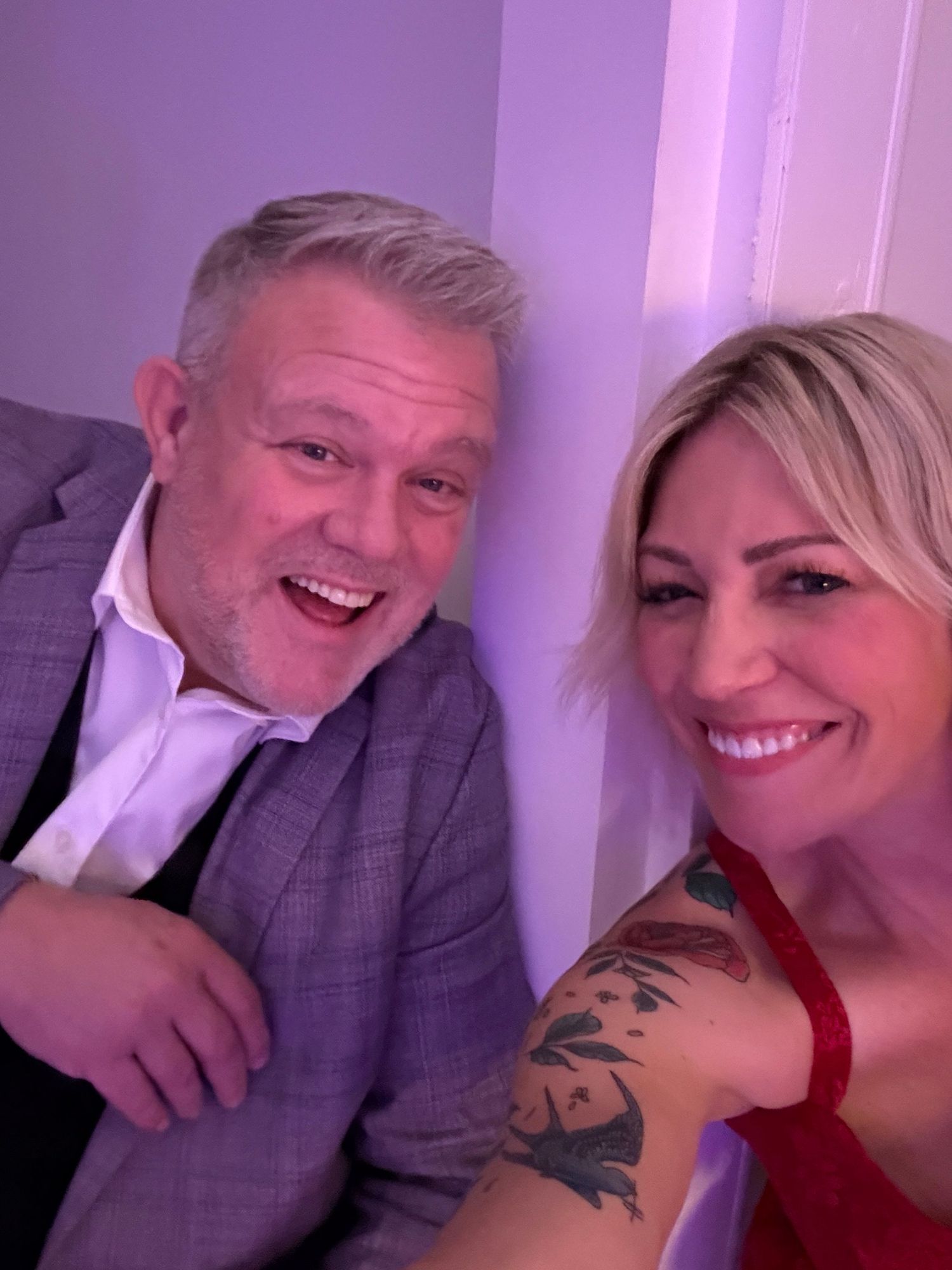 A couple in a purple-lit room taking playful selfies together, with one wearing a gray suit and the other in red.