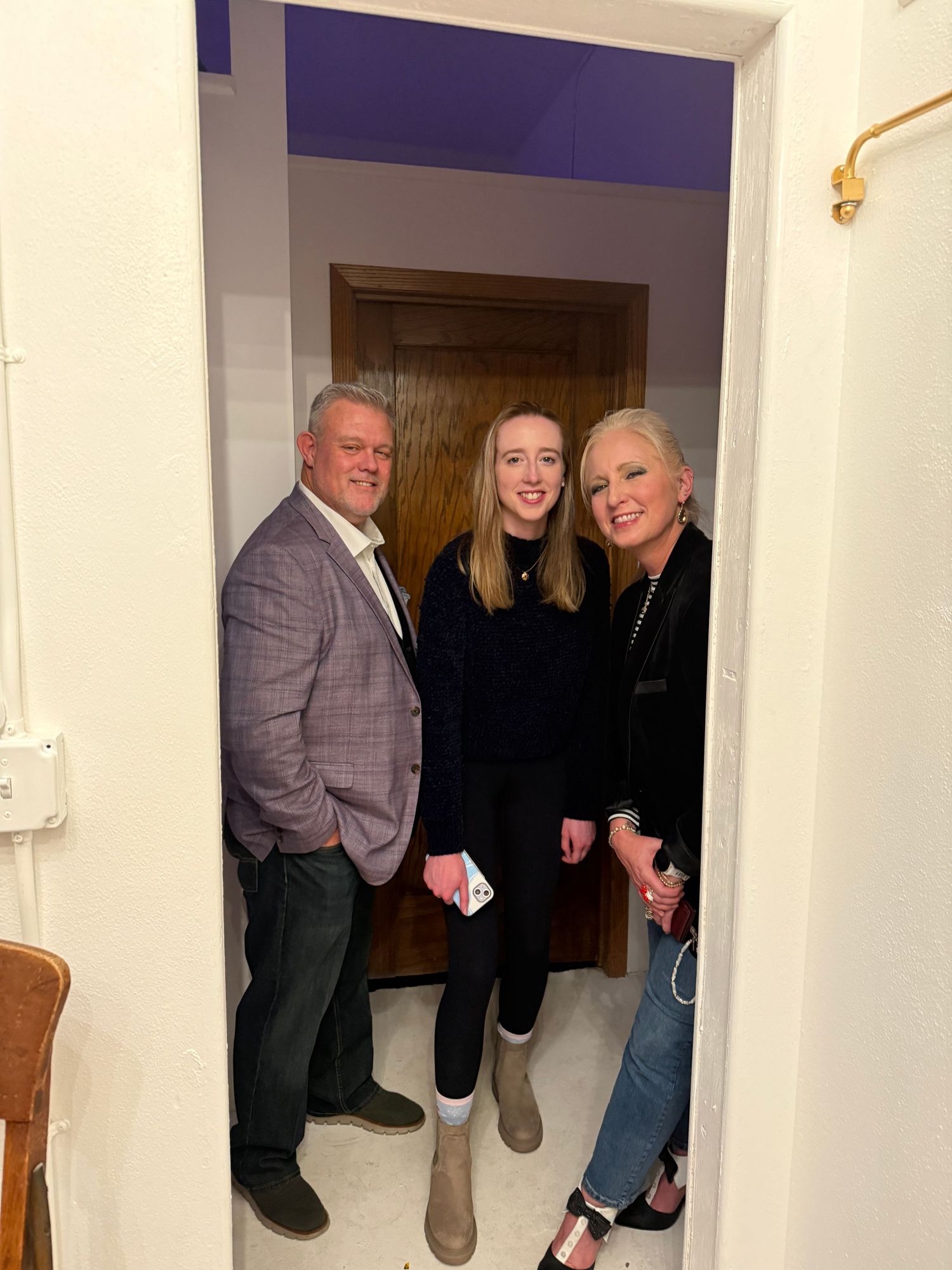 Three people stand together smiling in a doorway of a home with white walls and a dark wooden door.