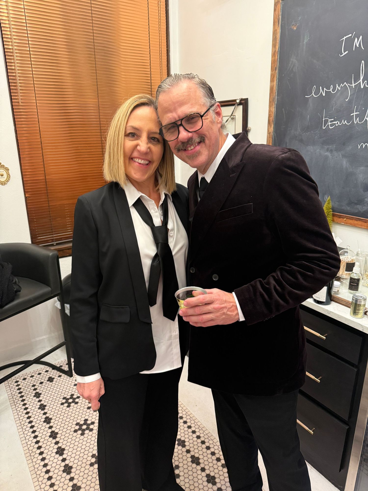 Two people in formal business attire share a light moment in an office setting with a chalkboard wall in the background.