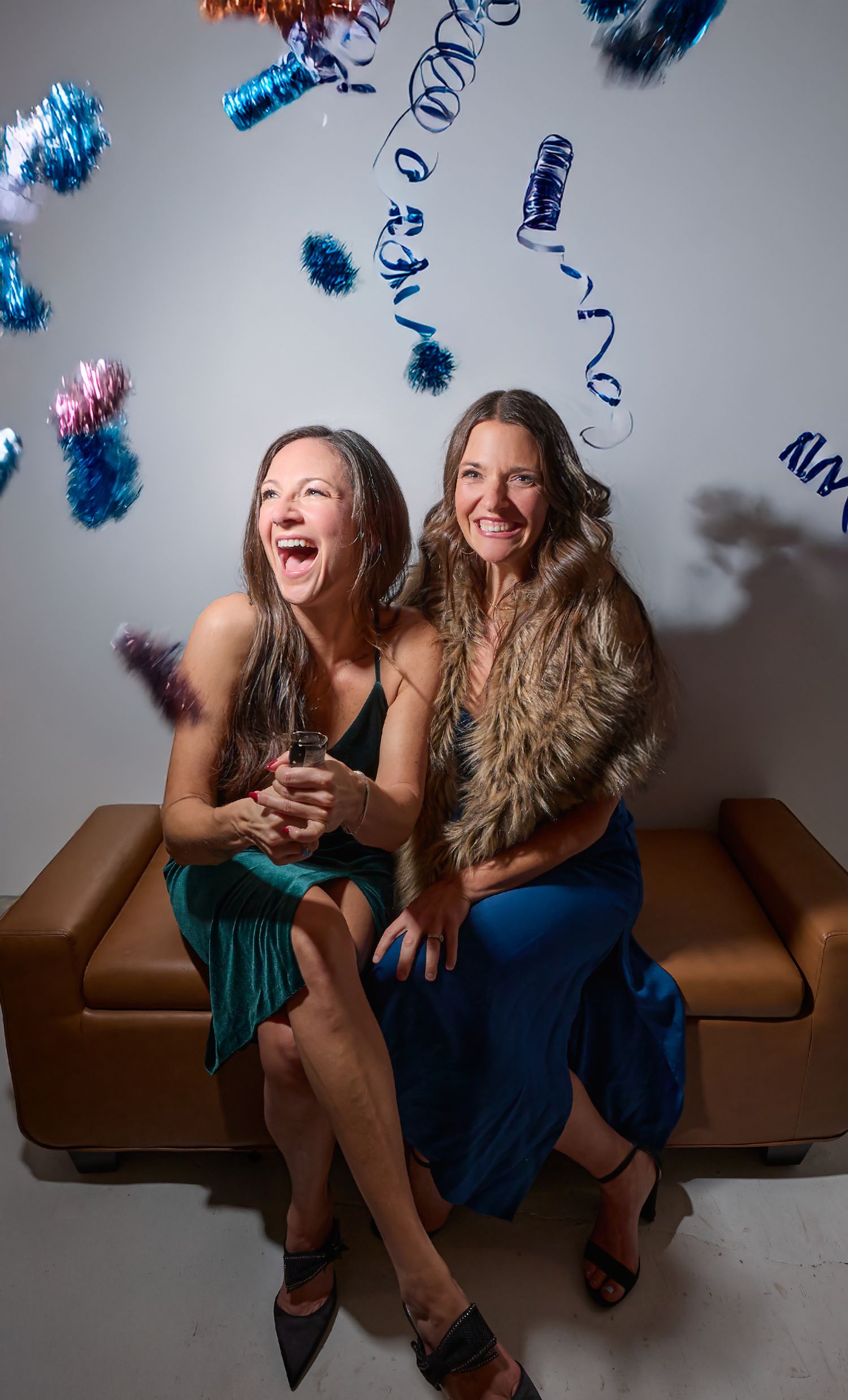 Two people laughing together on a brown leather couch while blue and pink party streamers fall from above.