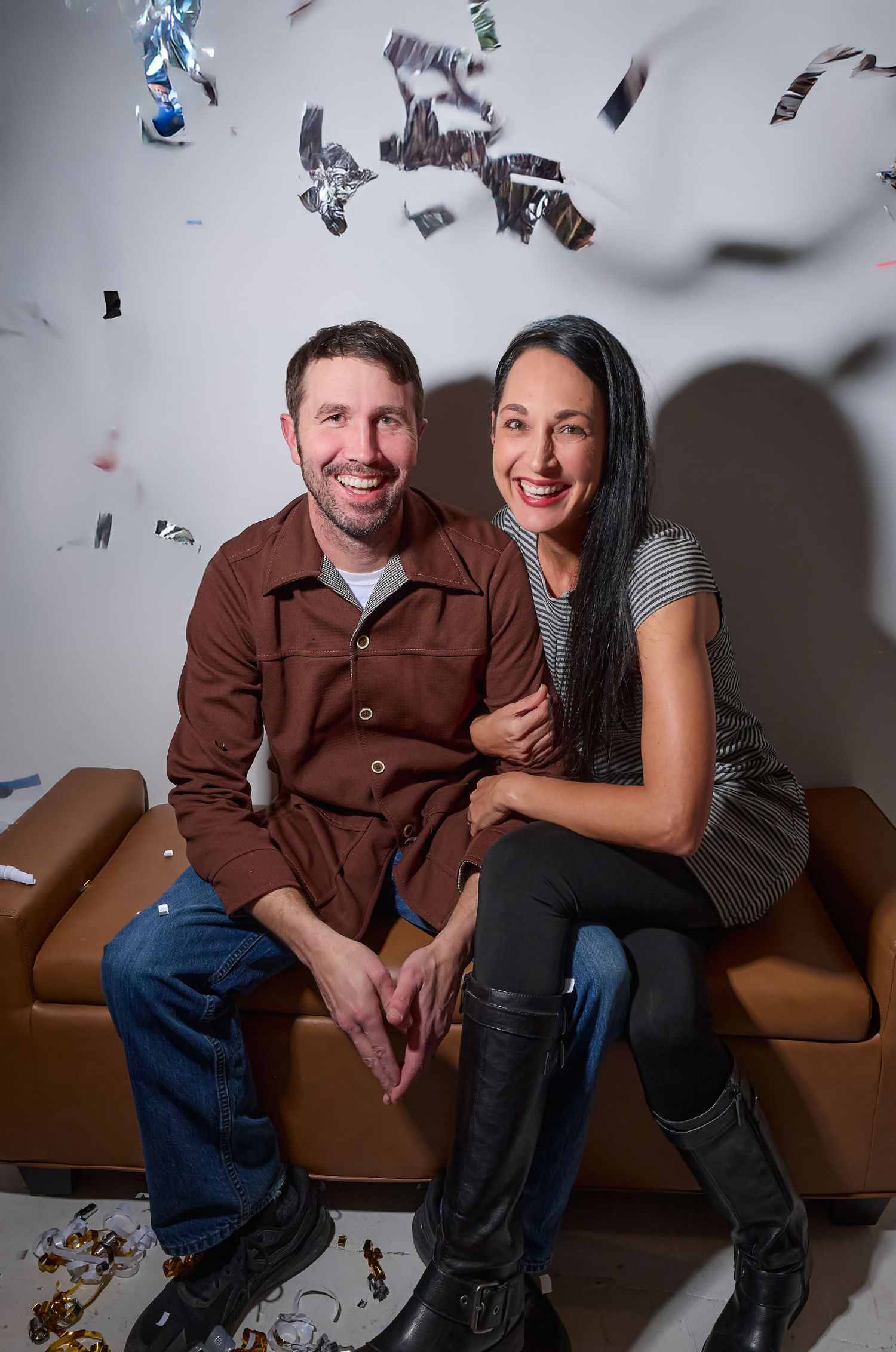 Two people sitting close together on a brown leather couch with silver confetti falling in the background.