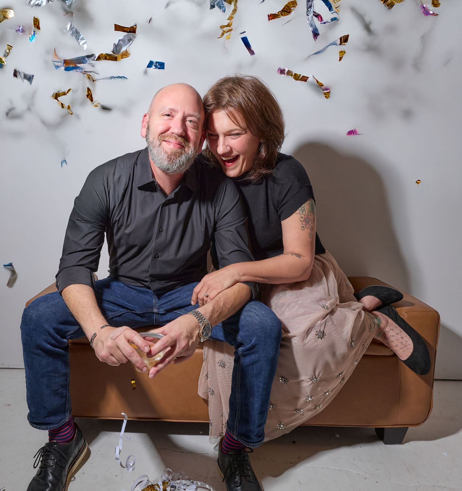 Two people sharing a joyful moment on a brown leather couch with colorful confetti raining down.