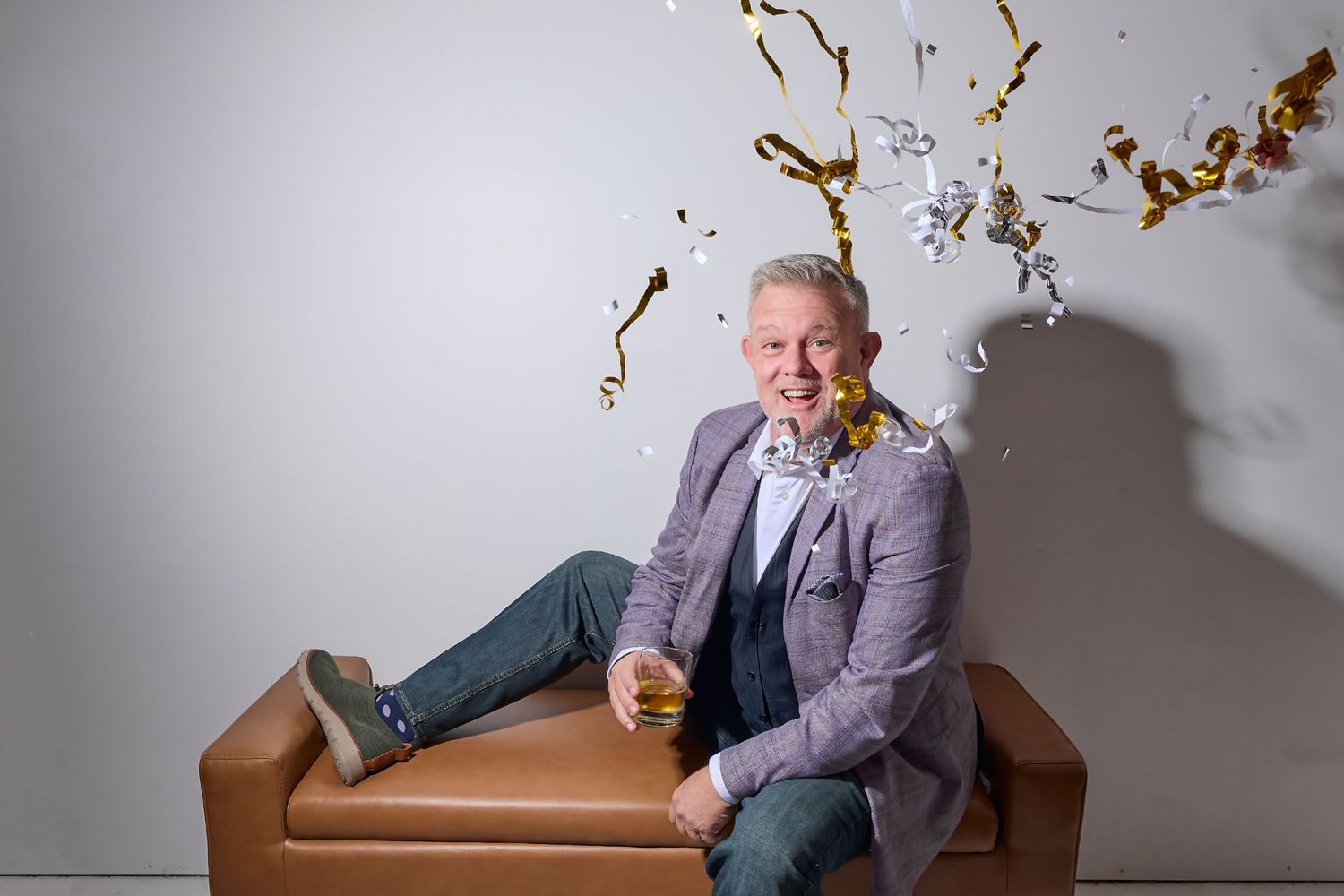 Person in gray blazer sitting on brown leather couch while autumn leaves fall in studio setting.
