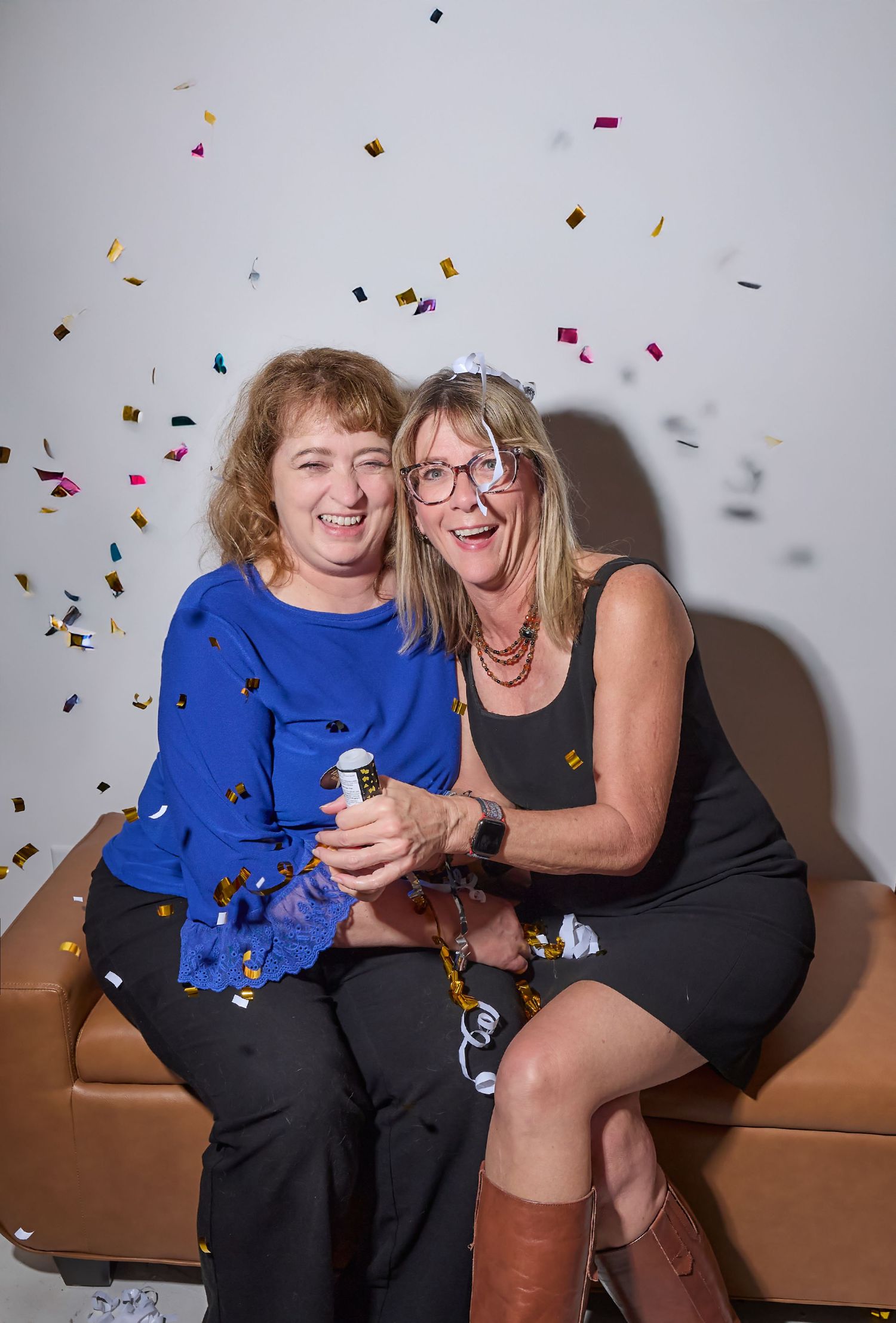 Two people sitting on a brown couch share joyful laughter as colorful confetti falls around them in a celebratory moment.