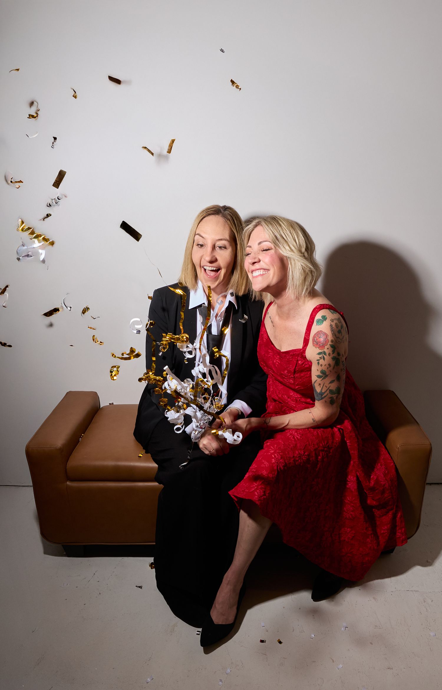 Two friends in formal attire laugh and celebrate with gold confetti on a brown couch during a festive photoshoot.