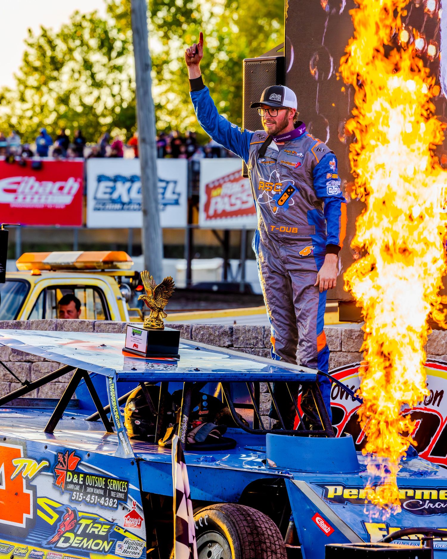 Race car driver celebrates victory atop blue stock car with dramatic fire pyrotechnics at motorsports event.