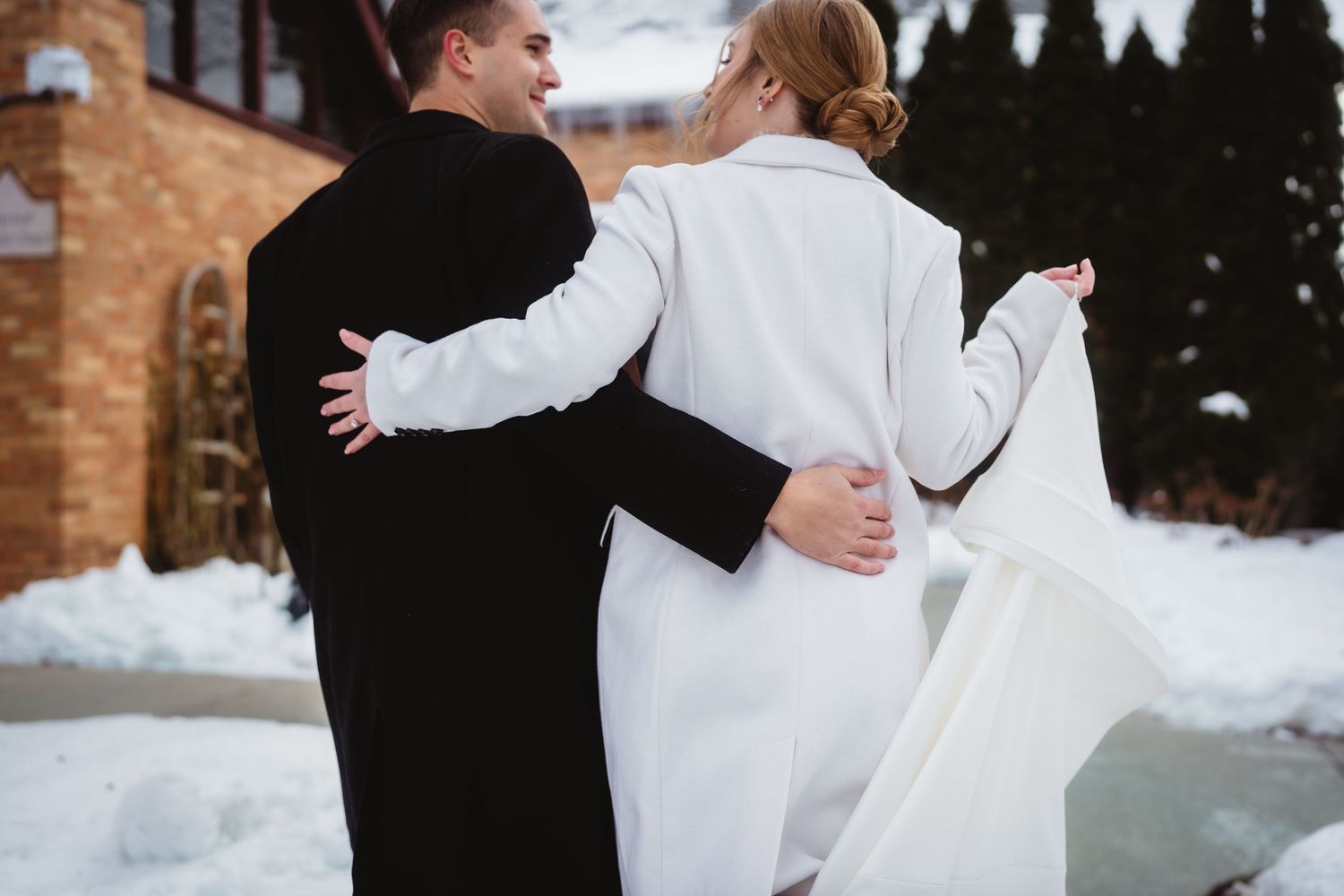 A couple in winter wedding attire embrace and dance together in the snow against a rustic brick backdrop.
