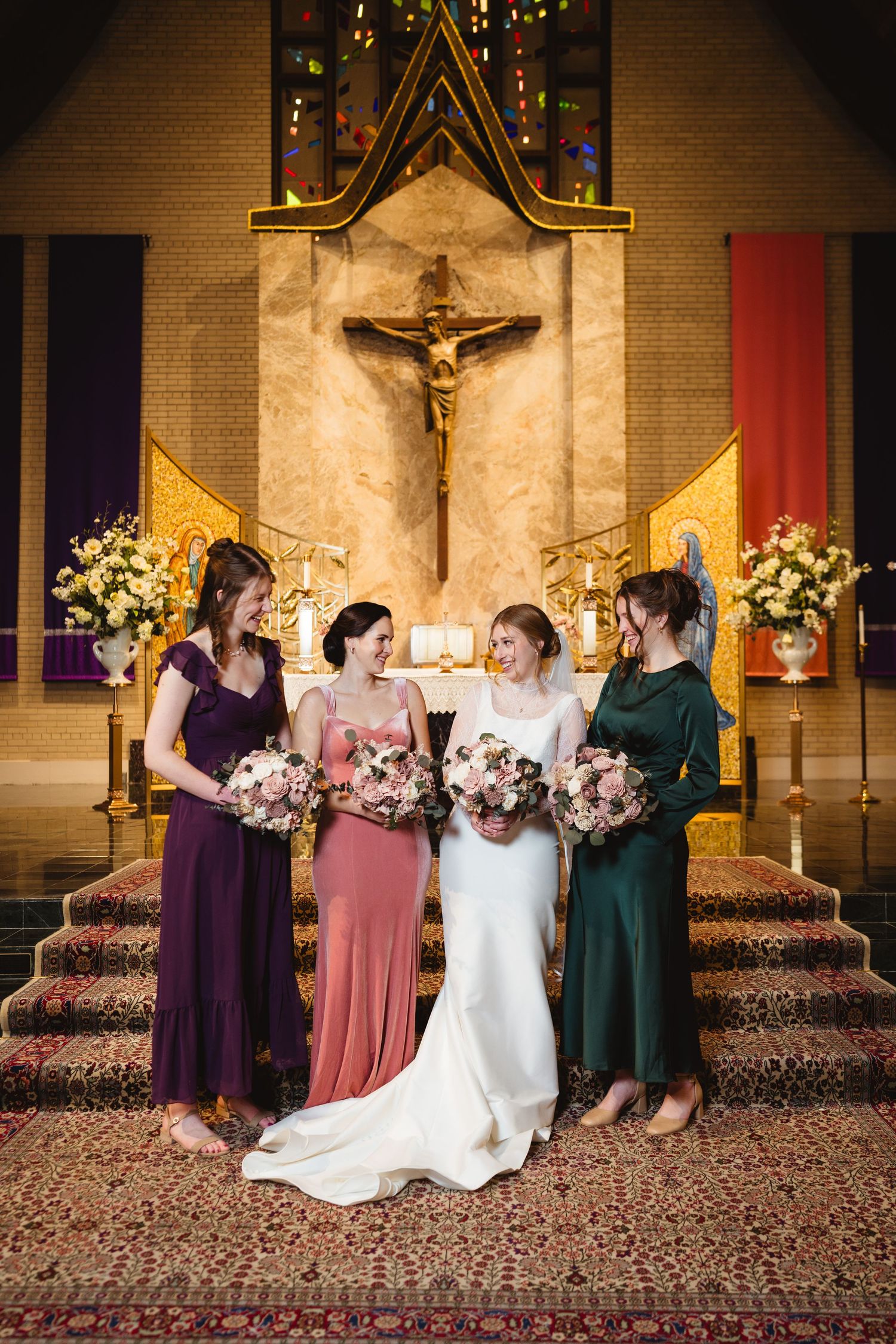 Wedding party in jewel-toned dresses pose at church altar with crucifix and floral arrangements in background.