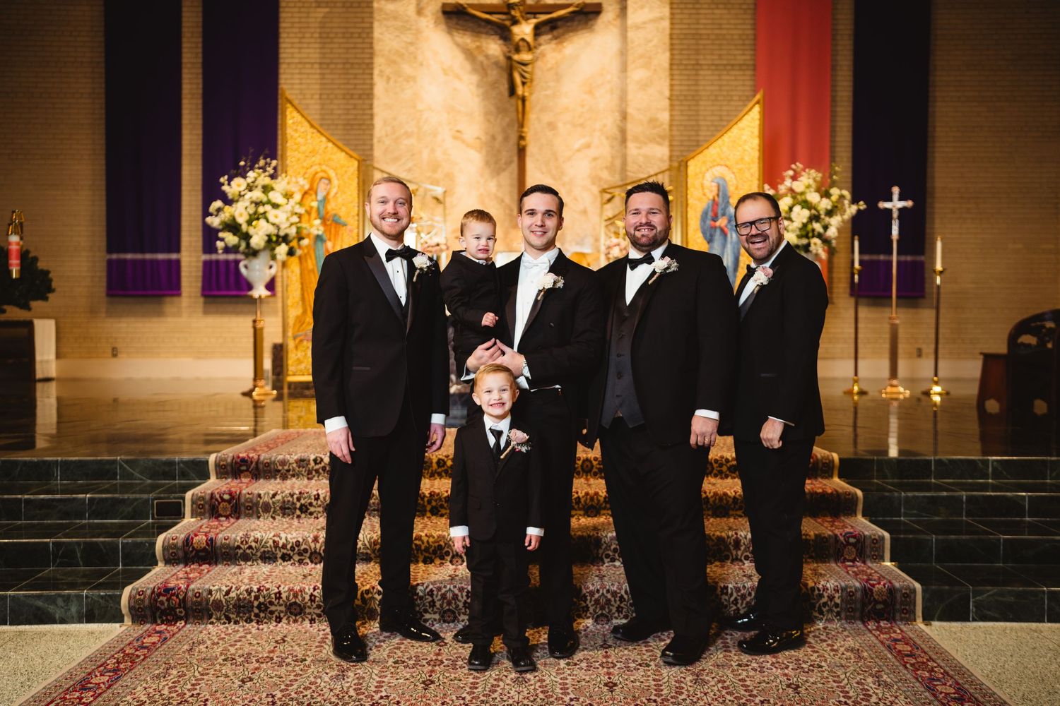 Wedding party in formal black attire stands in a line against a church backdrop with warm lighting and religious decor.